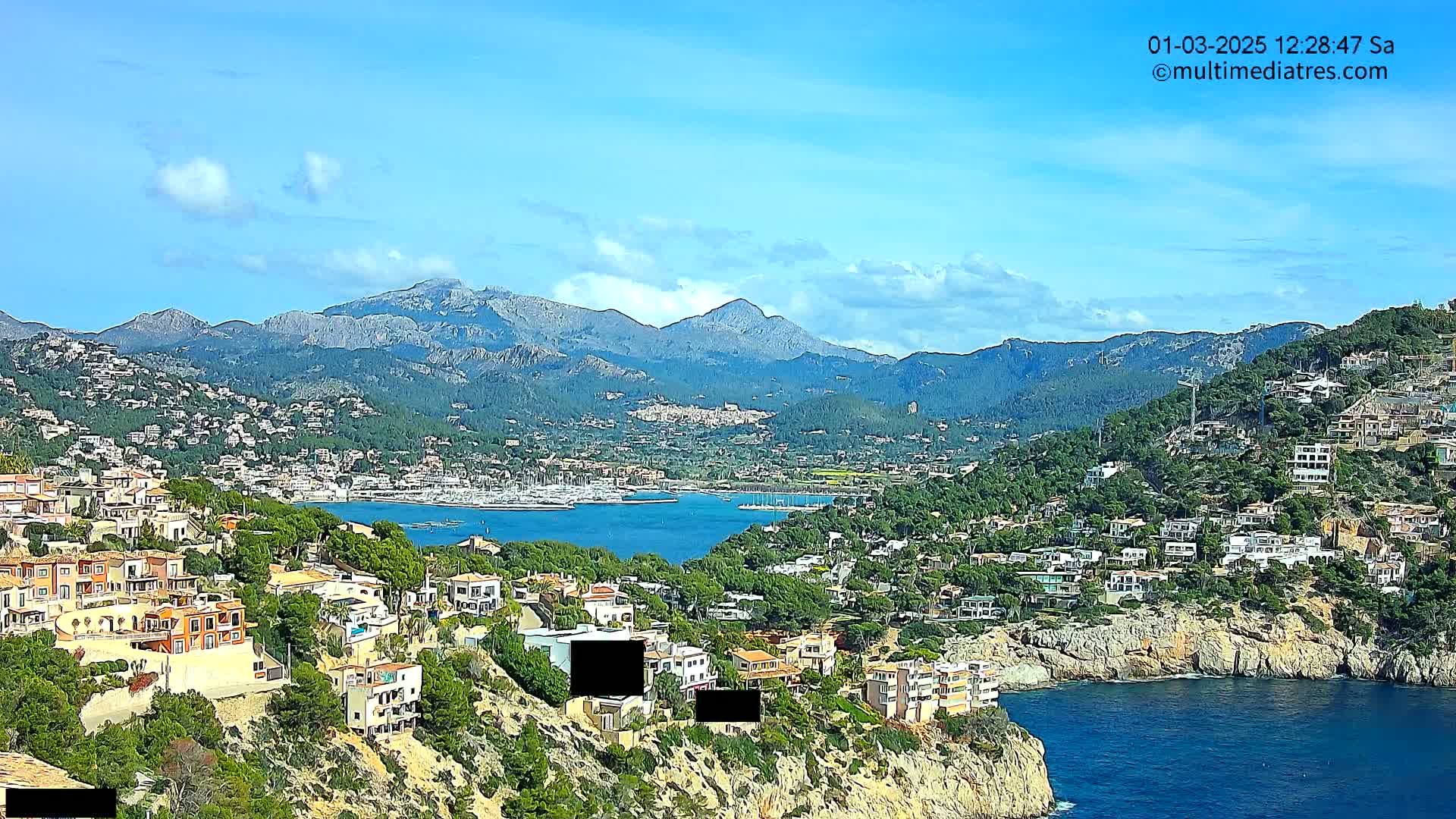 A coastal town nestled against a mountain range under a mostly sunny sky, with a calm body of water separating the town from a marina.