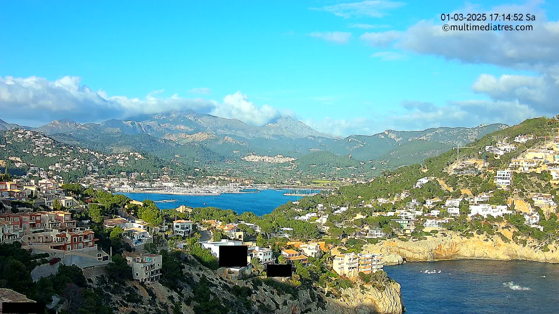 A coastal town nestled against a mountainous backdrop under a partly cloudy sky, featuring a calm bay with numerous boats.