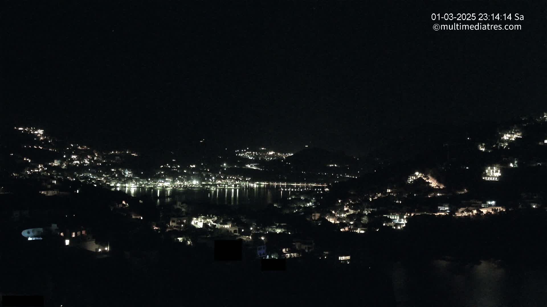 A nighttime view of a coastal town nestled on hillsides, with its lights illuminating the dark landscape and reflecting on a calm body of water.