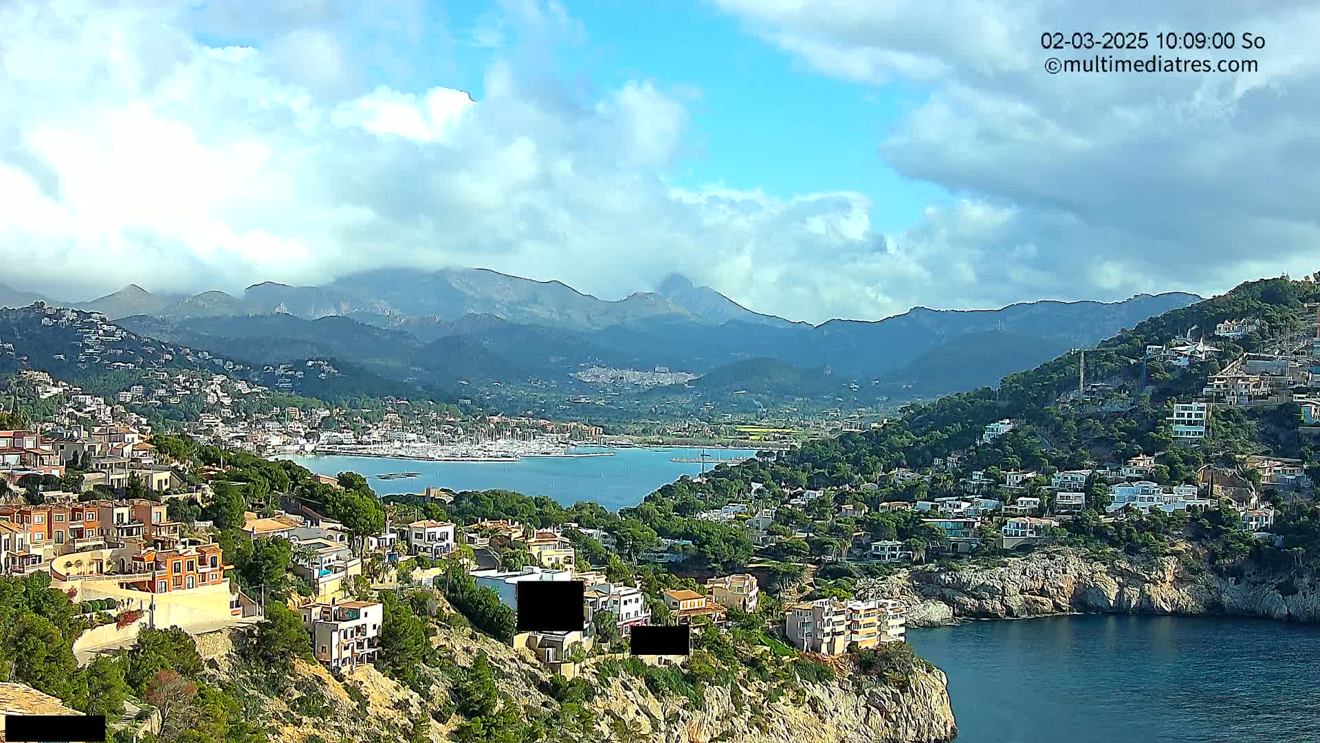 A coastal town nestled in hills overlooking a calm bay filled with boats, with mountains in the background under a partly cloudy sky.