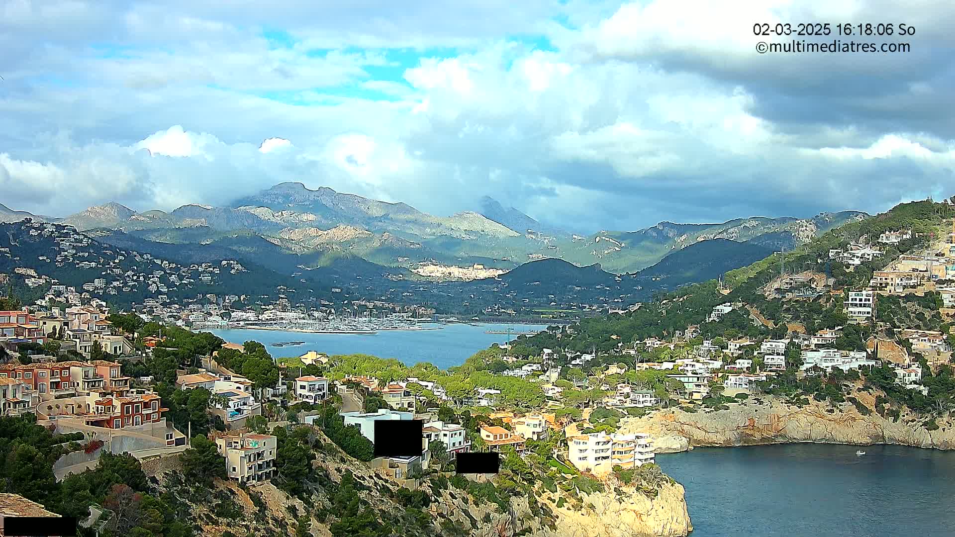 A coastal town nestled in a mountainous region, under a partly cloudy sky, overlooks a calm bay filled with boats.