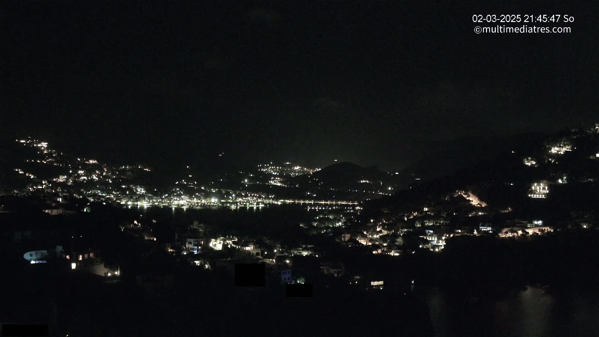 A nighttime view shows a coastal town nestled in hills, with many lights illuminating the buildings and streets.