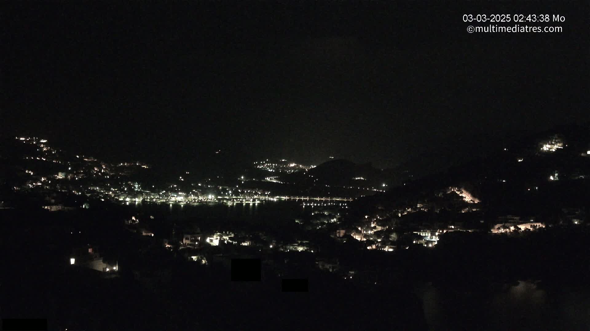 A nighttime view of a coastal town nestled on hillsides, with scattered lights illuminating the landscape under a clear, dark sky.