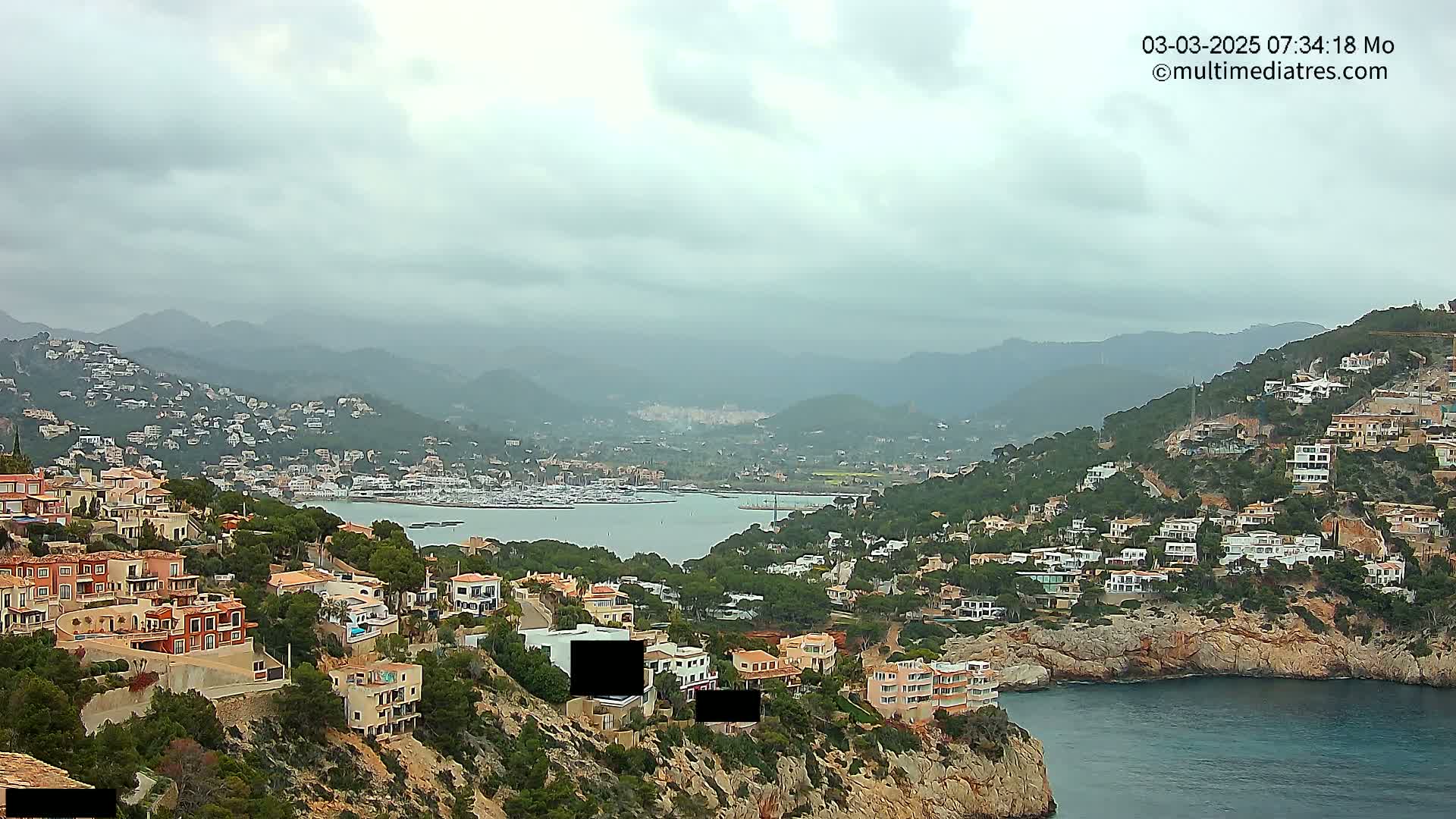 A coastal town nestled in hills overlooking a calm bay filled with boats under an overcast sky.