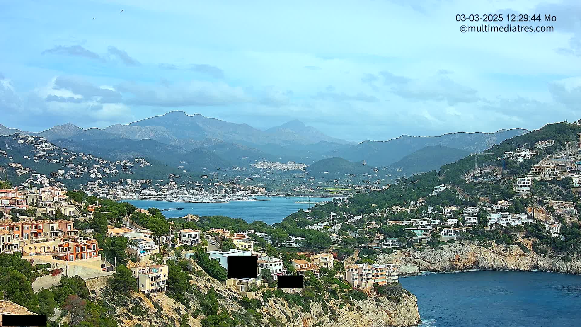 A coastal town nestled on a hillside overlooking a calm bay, with mountains in the background under a partly cloudy sky.