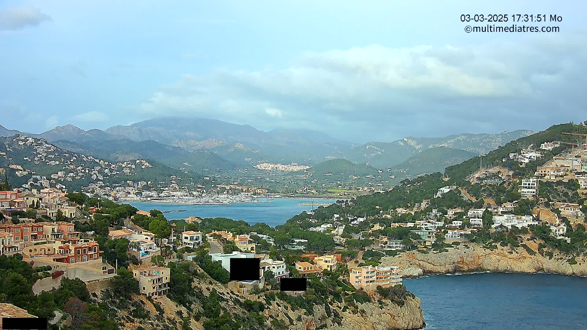 A coastal town nestled on a hillside overlooking a calm bay, with mountains in the background under a partly cloudy sky.