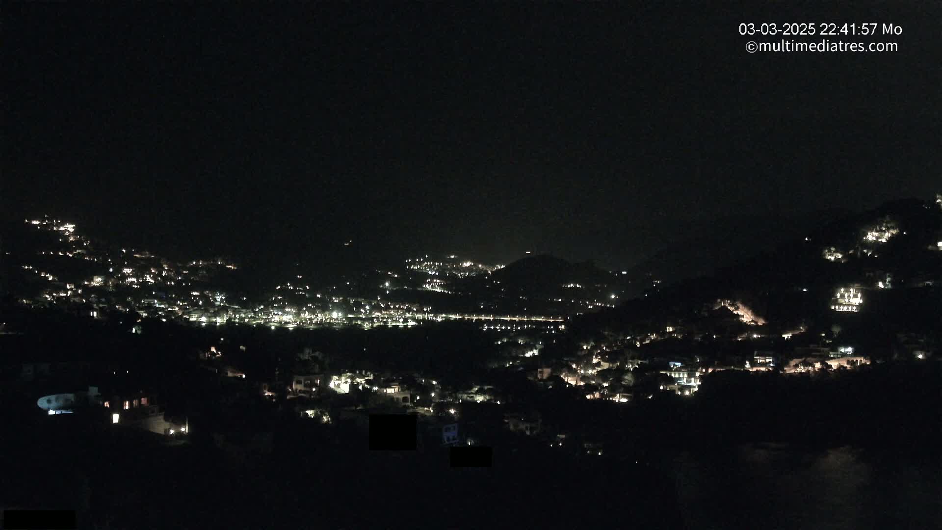 A nighttime, long-range view shows a city nestled in dark hills, illuminated by scattered house lights.