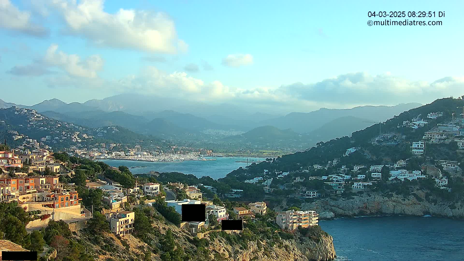 A coastal town nestled on a hillside overlooks a calm bay filled with boats, all set against a backdrop of hazy mountains under a partly cloudy sky.