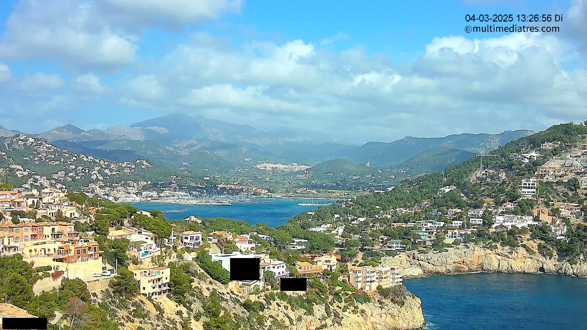 A partly cloudy day overlooking a coastal town nestled between hills and a calm bay filled with boats, mountains visible in the distance.