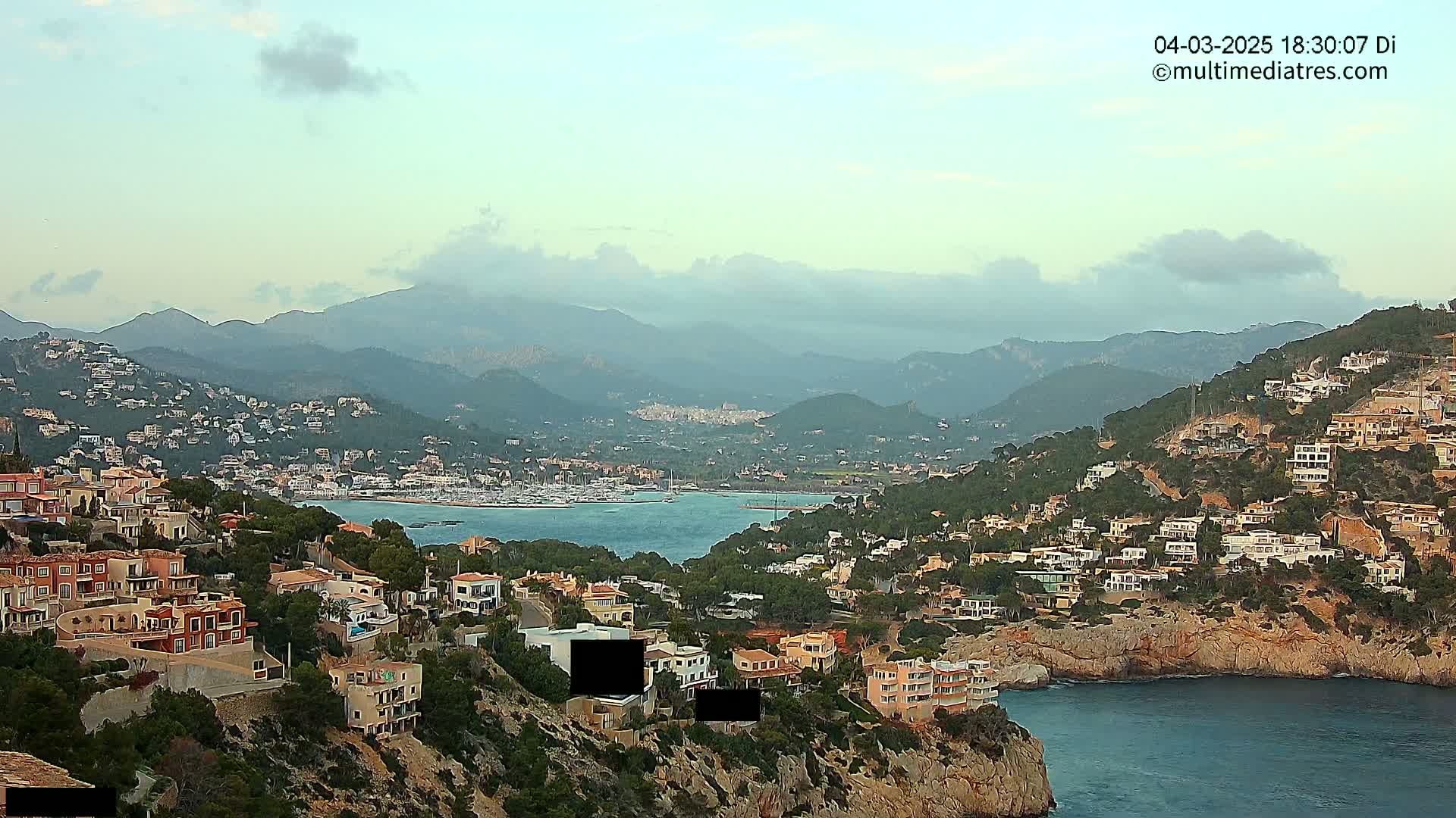 A coastal town with numerous houses clinging to hillsides overlooking a calm bay and distant mountains under a partly cloudy sky.