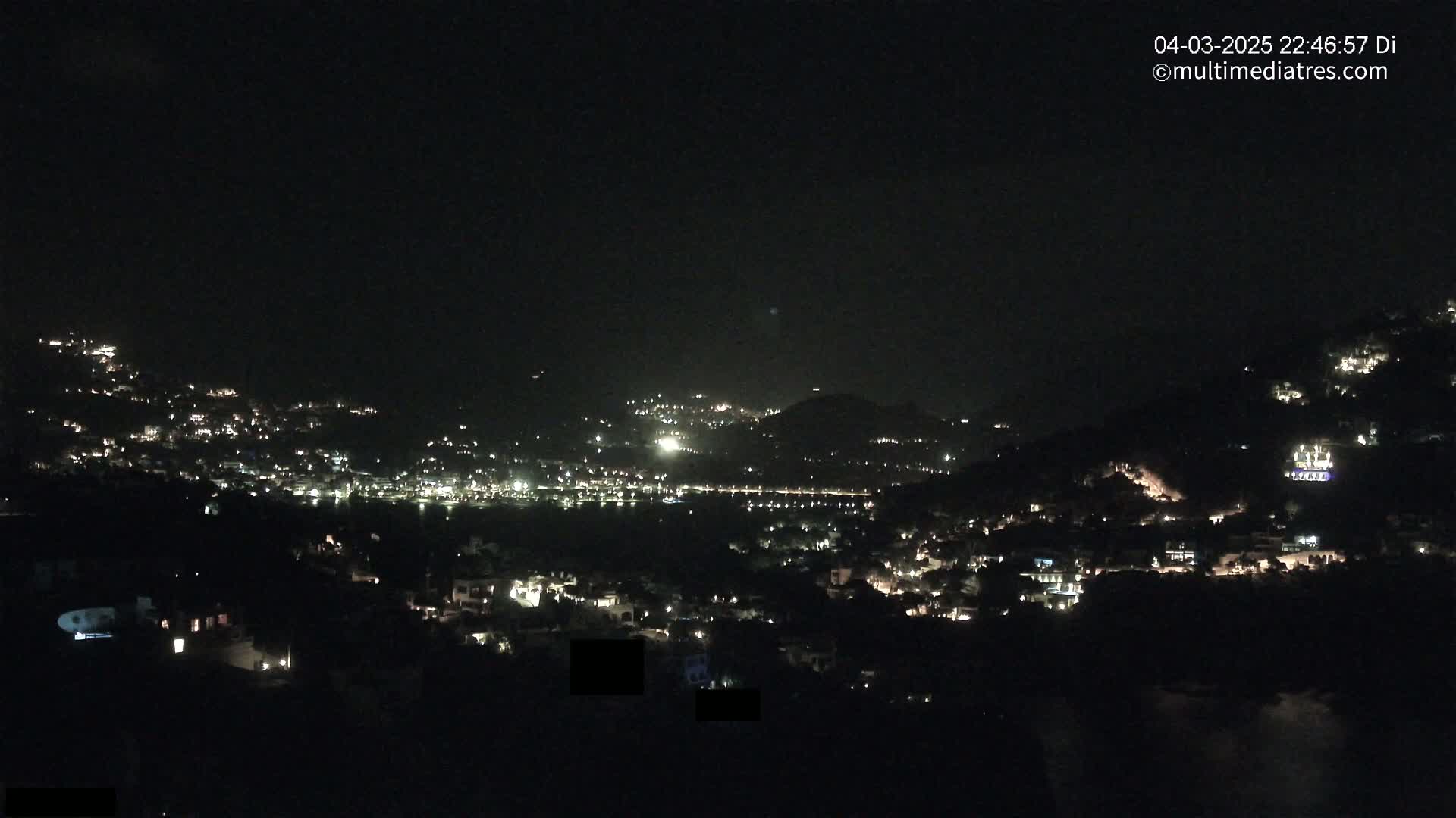 A nighttime view of a hillside town with scattered lights, overlooking a dark body of water under a clear, dark sky.