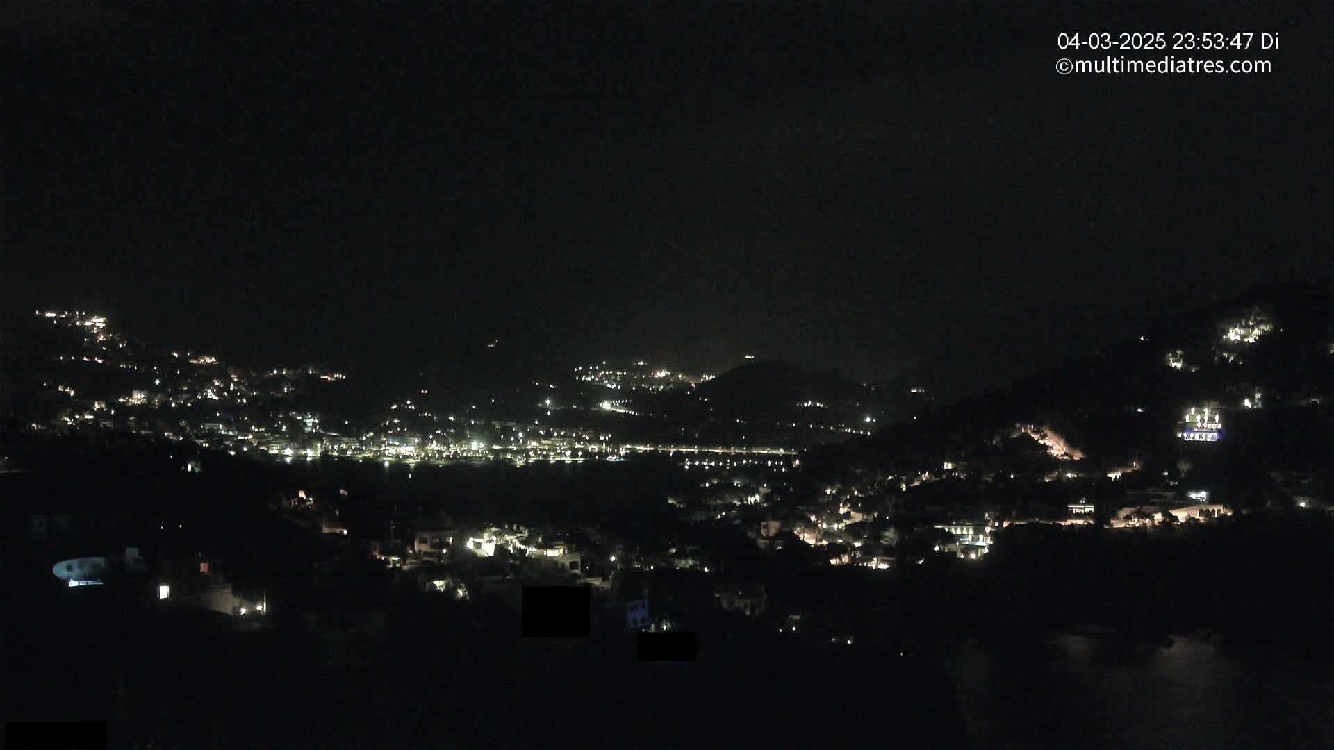 A nighttime view shows a hillside town illuminated by scattered lights under a clear, dark sky.
