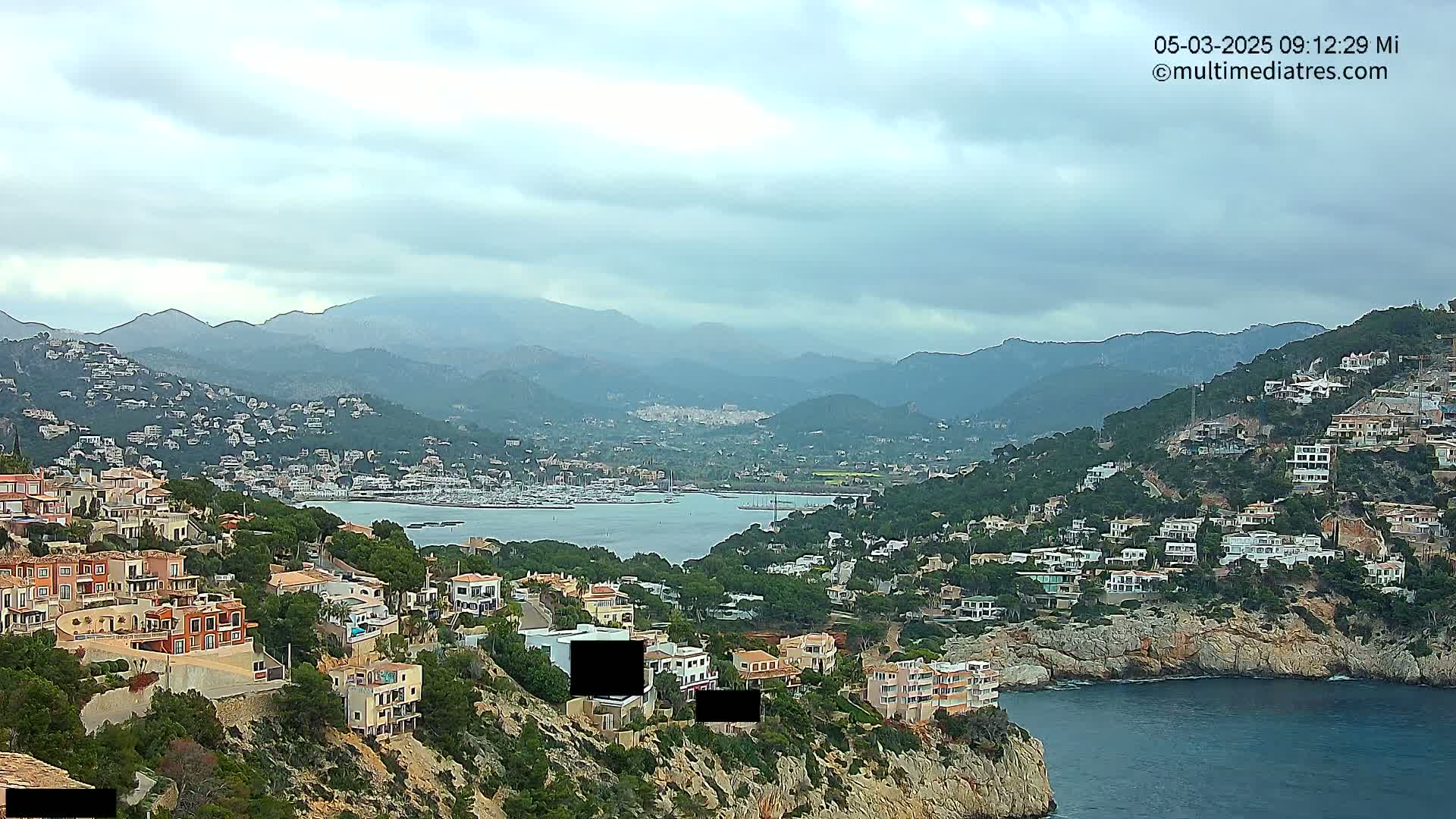 A coastal town nestled in hills overlooking a calm bay filled with boats under a cloudy sky.