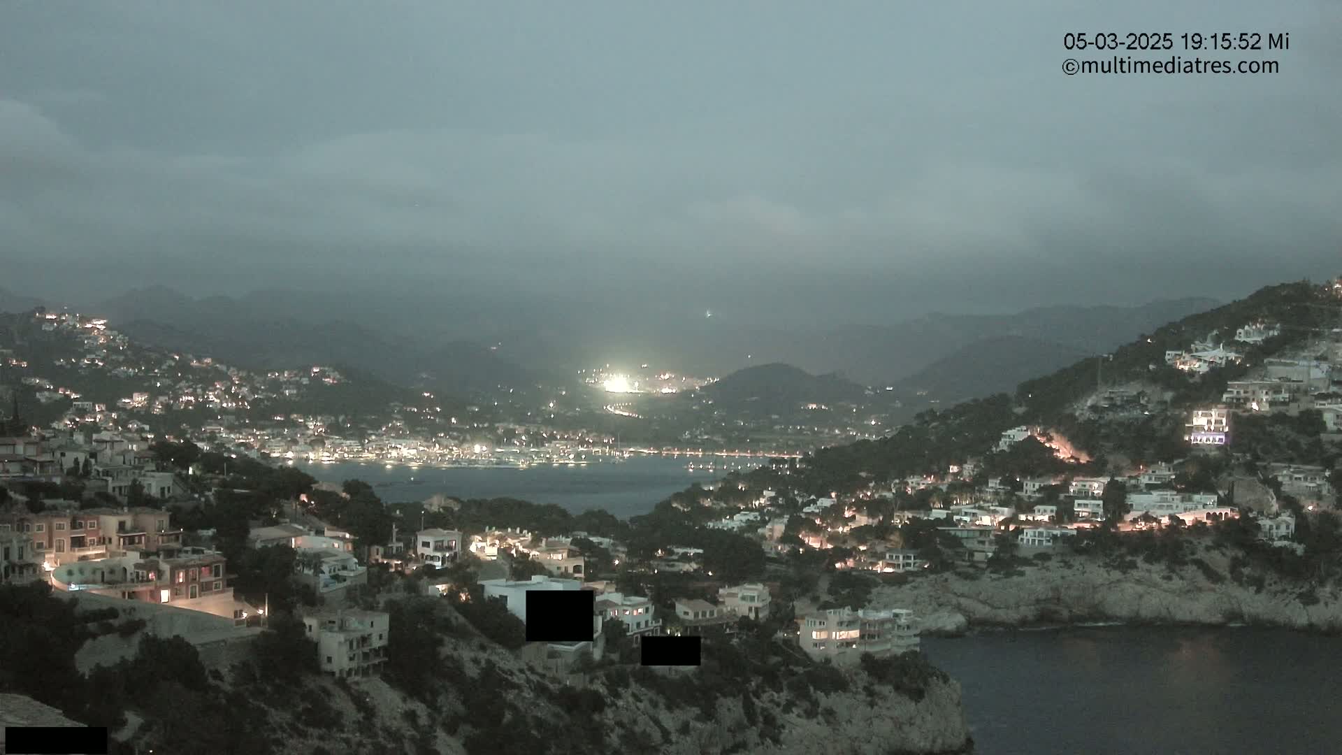A coastal town at twilight under a cloudy sky is nestled among hills, with the lights of buildings illuminating the landscape.