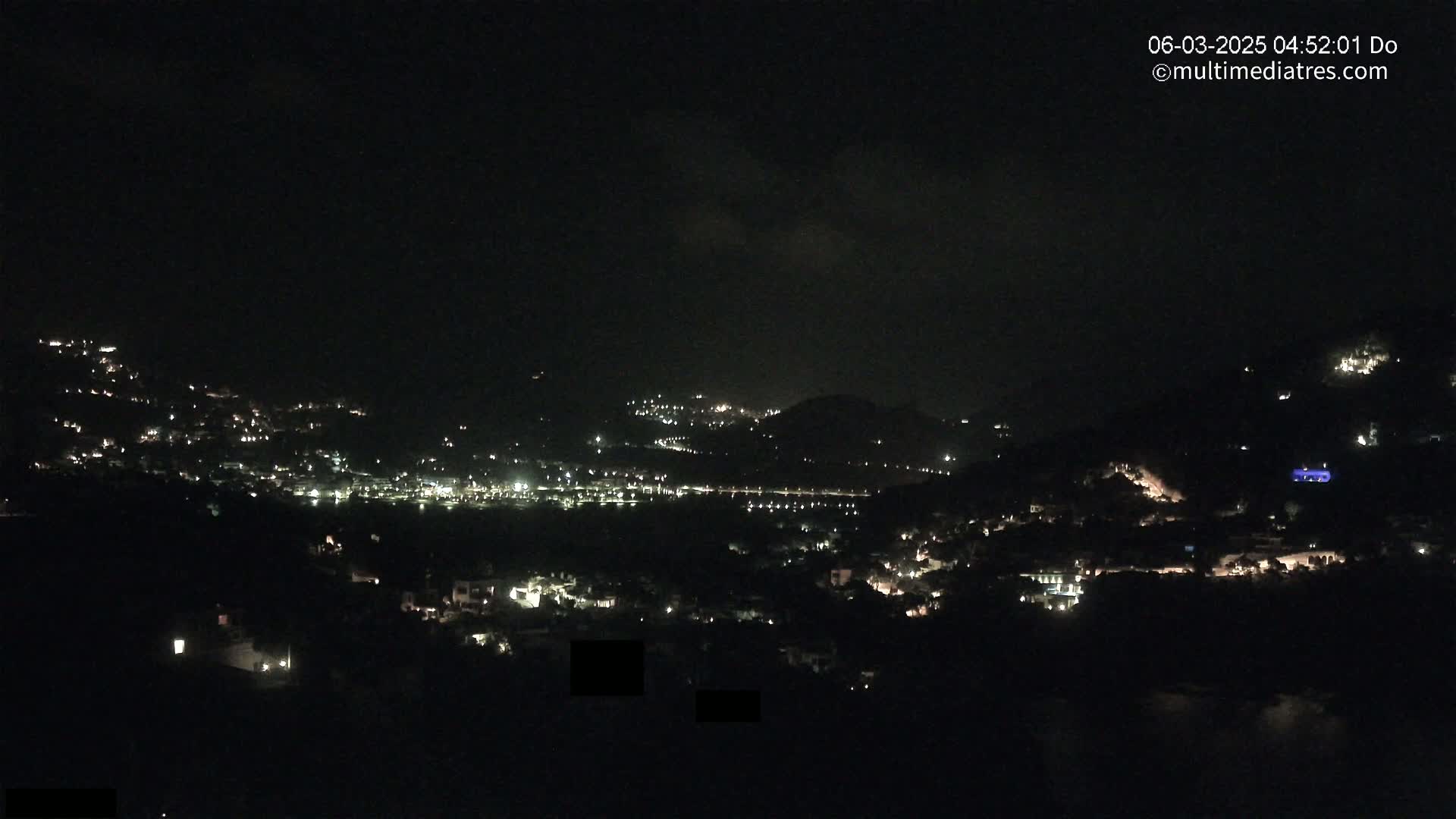 A nighttime view of a hilly town with scattered lights under a mostly cloudy sky.