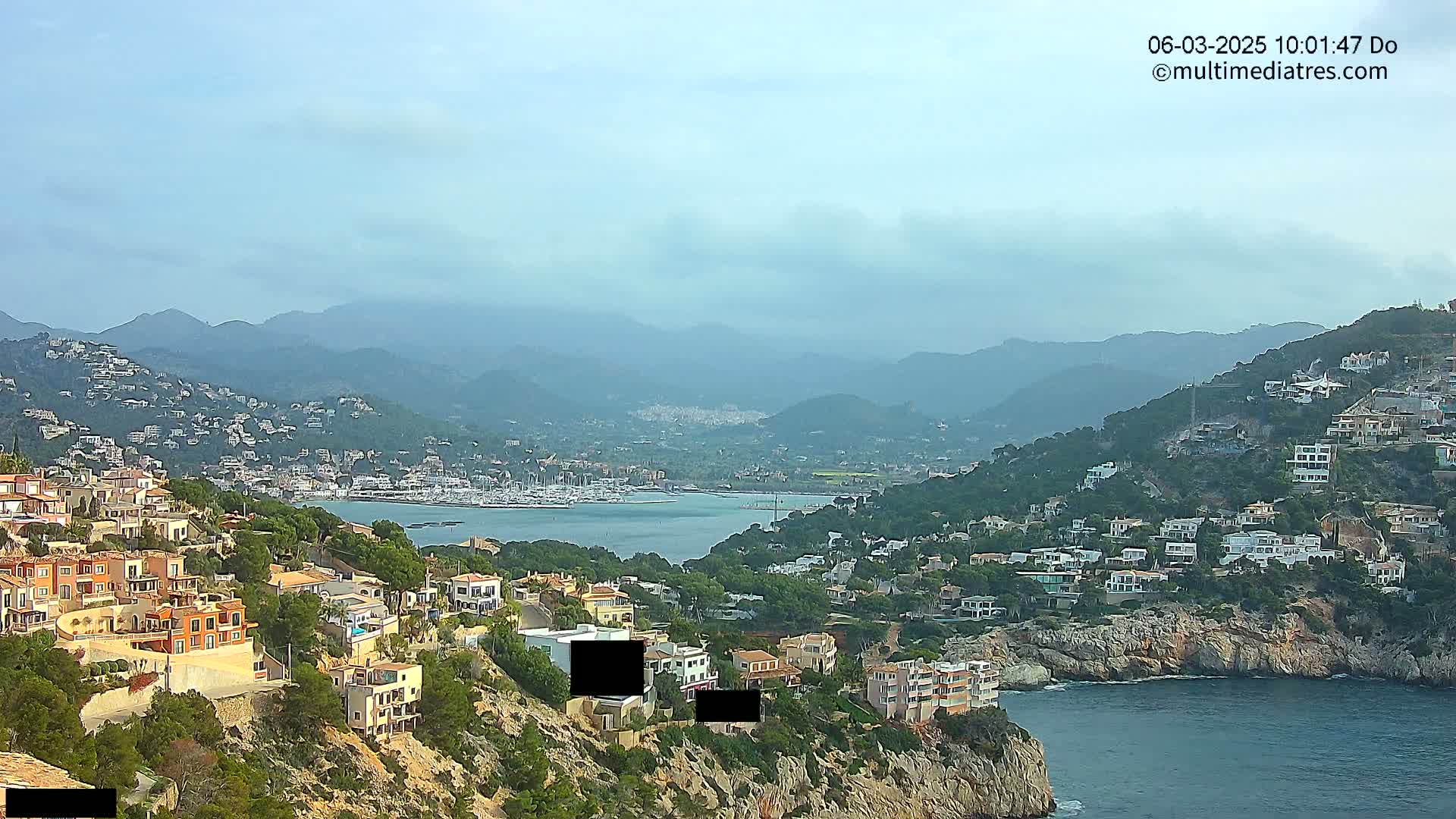 A coastal town nestled on a hillside overlooks a calm bay filled with boats, with hazy mountains in the background under a mostly clear sky.