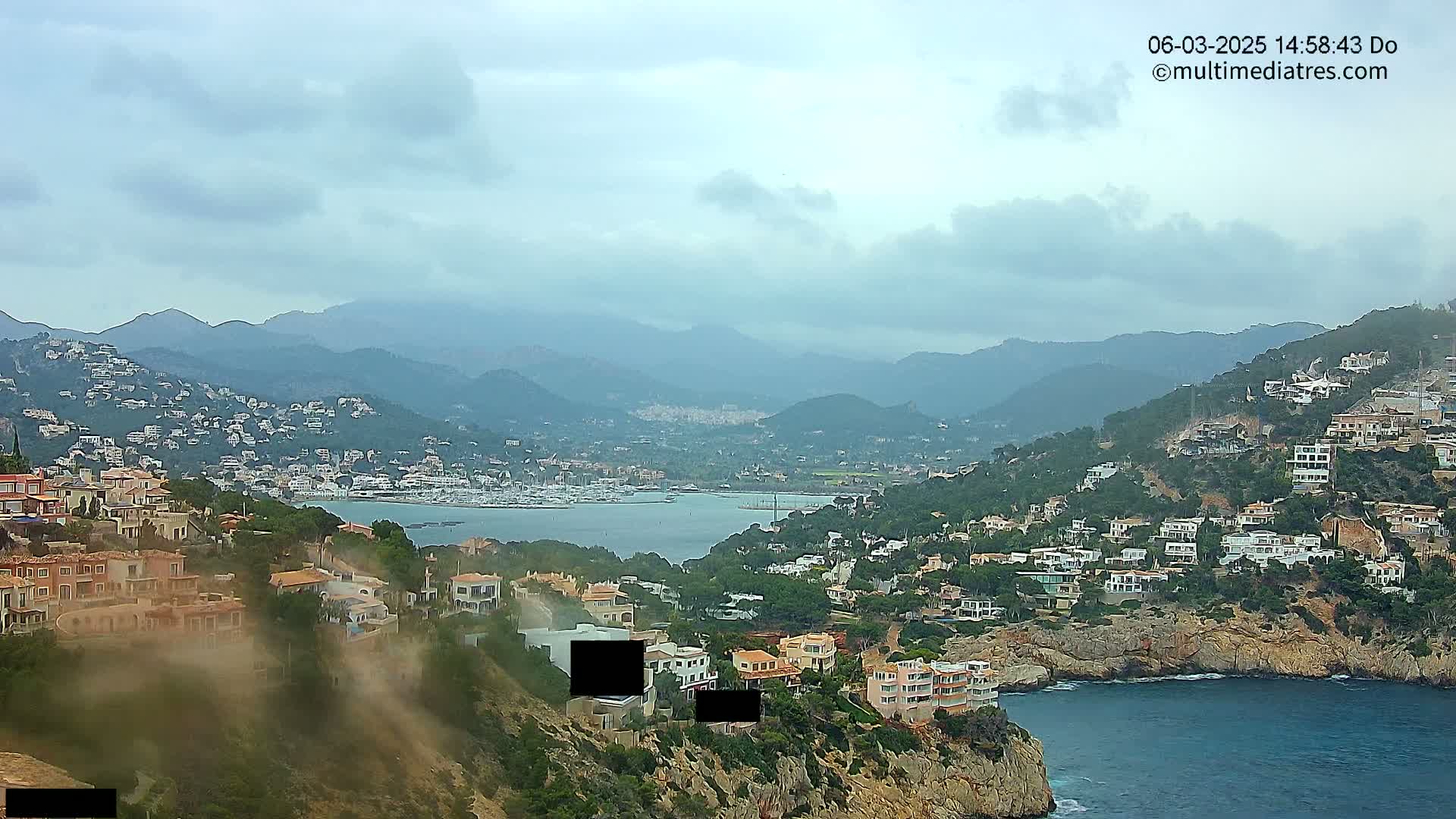 A coastal town nestled on a hillside overlooking a calm bay and a mountain range under a cloudy sky.