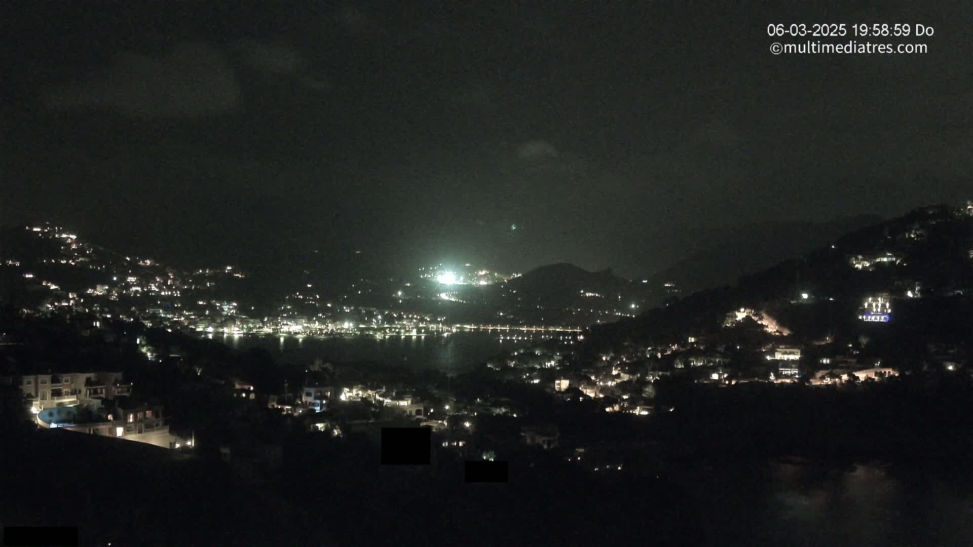 A nighttime view of a coastal town nestled on hillsides, with scattered lights illuminating the buildings and a dark body of water visible in the distance under a mostly clear sky.