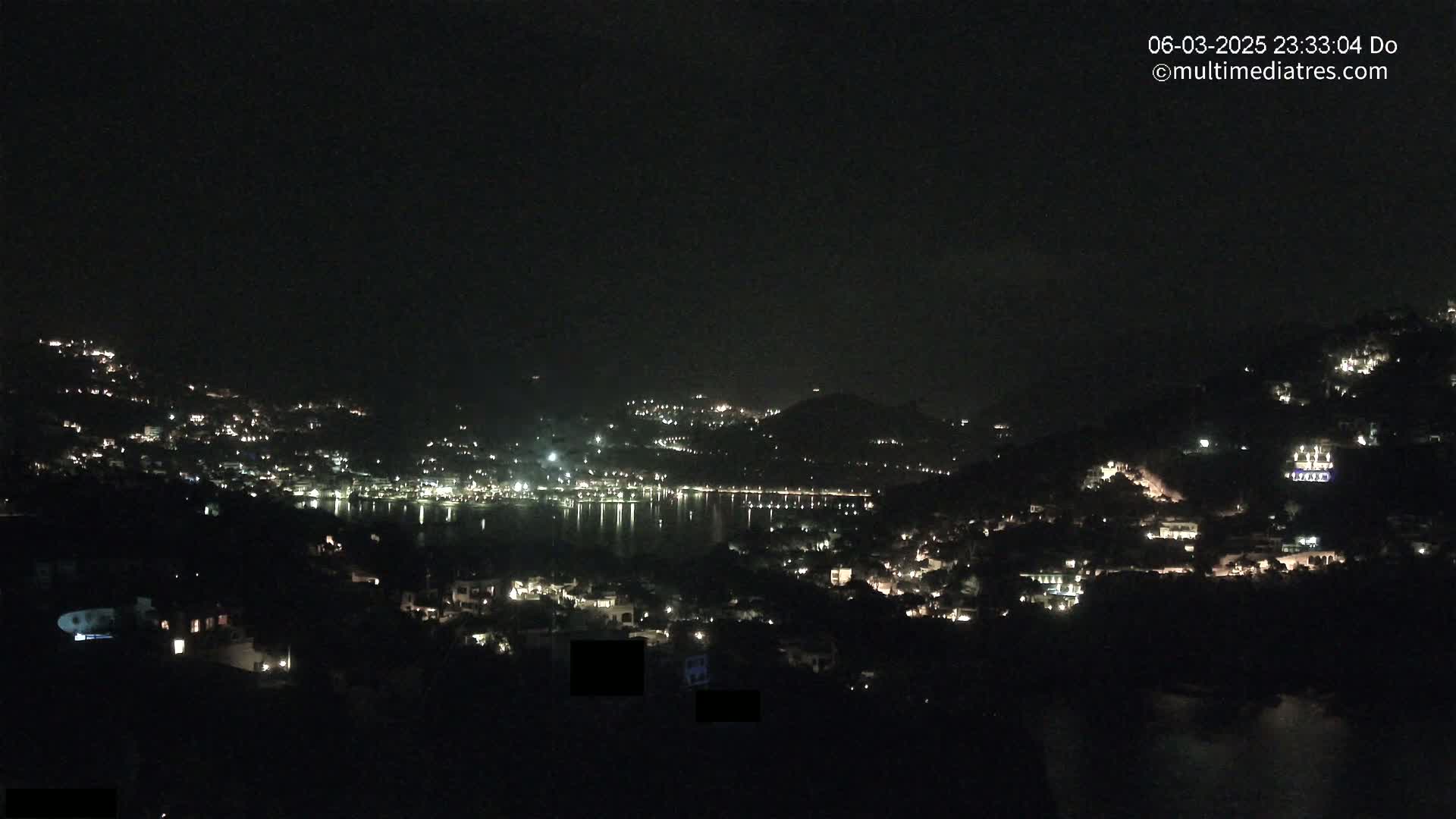 A nighttime view of a coastal town nestled on hillsides, with its lights reflecting on a calm body of water under a clear, dark sky.