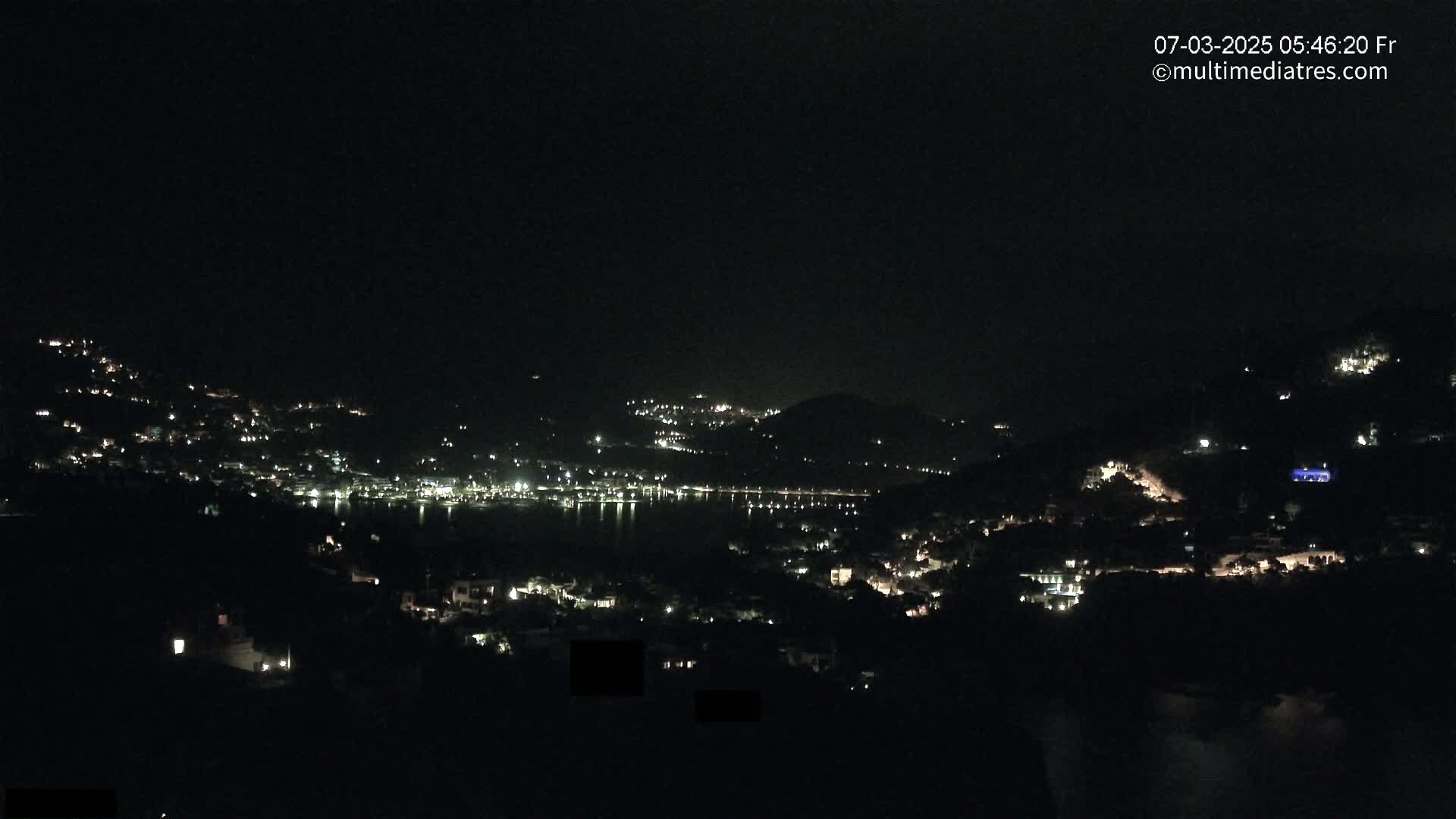 A nighttime view shows a coastal town with scattered lights illuminated against dark hills and a calm body of water.