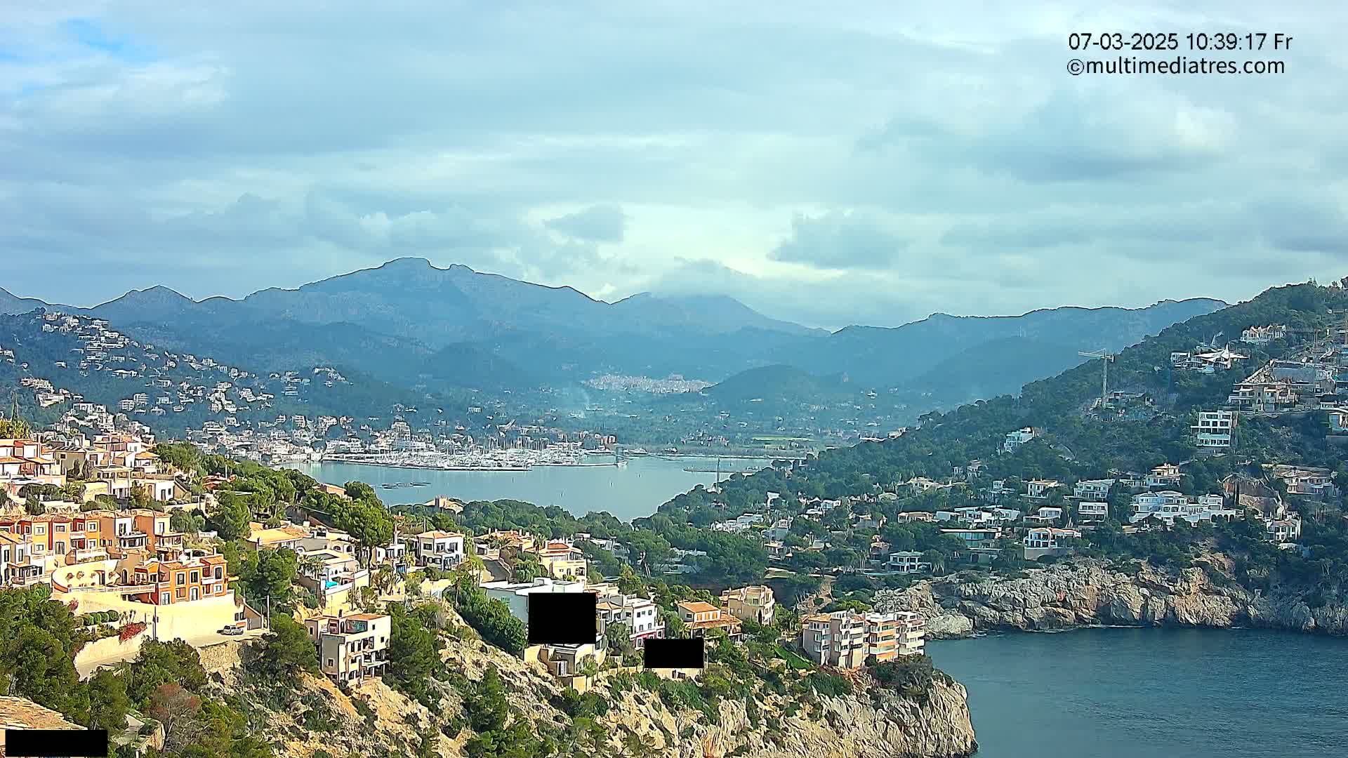 A coastal town nestled on a hillside overlooking a calm bay, with mountains in the background under a partly cloudy sky.