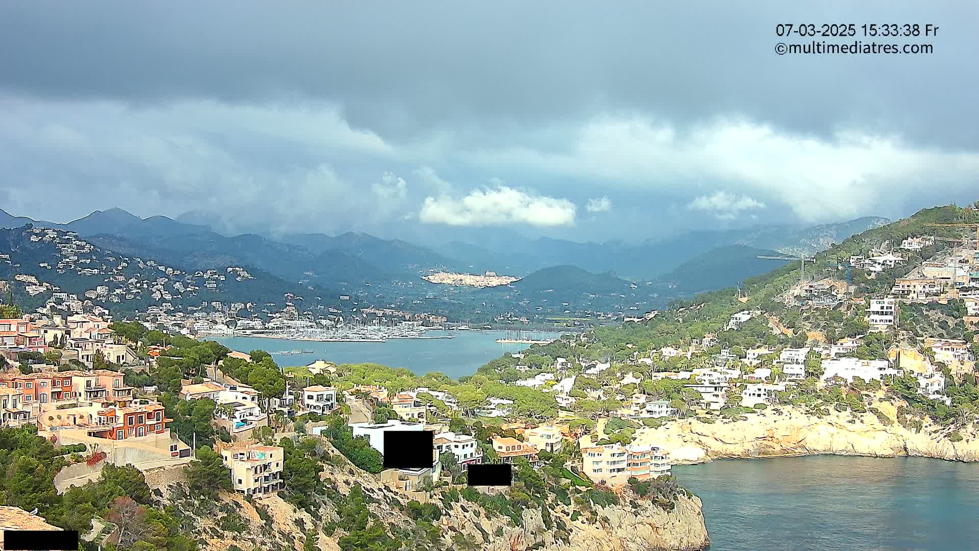 A coastal town nestled in a mountainous region under a cloudy sky overlooks a calm bay filled with boats.