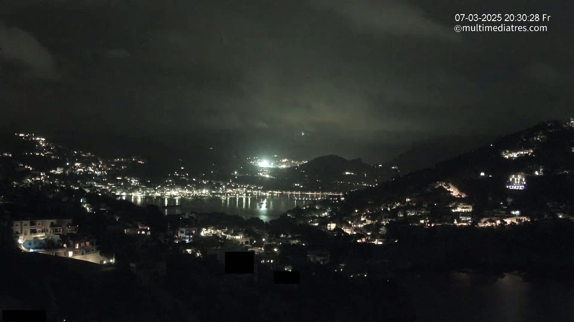 A nighttime view of a coastal town nestled on hillsides, with its lights reflecting on a calm body of water under a cloudy sky.