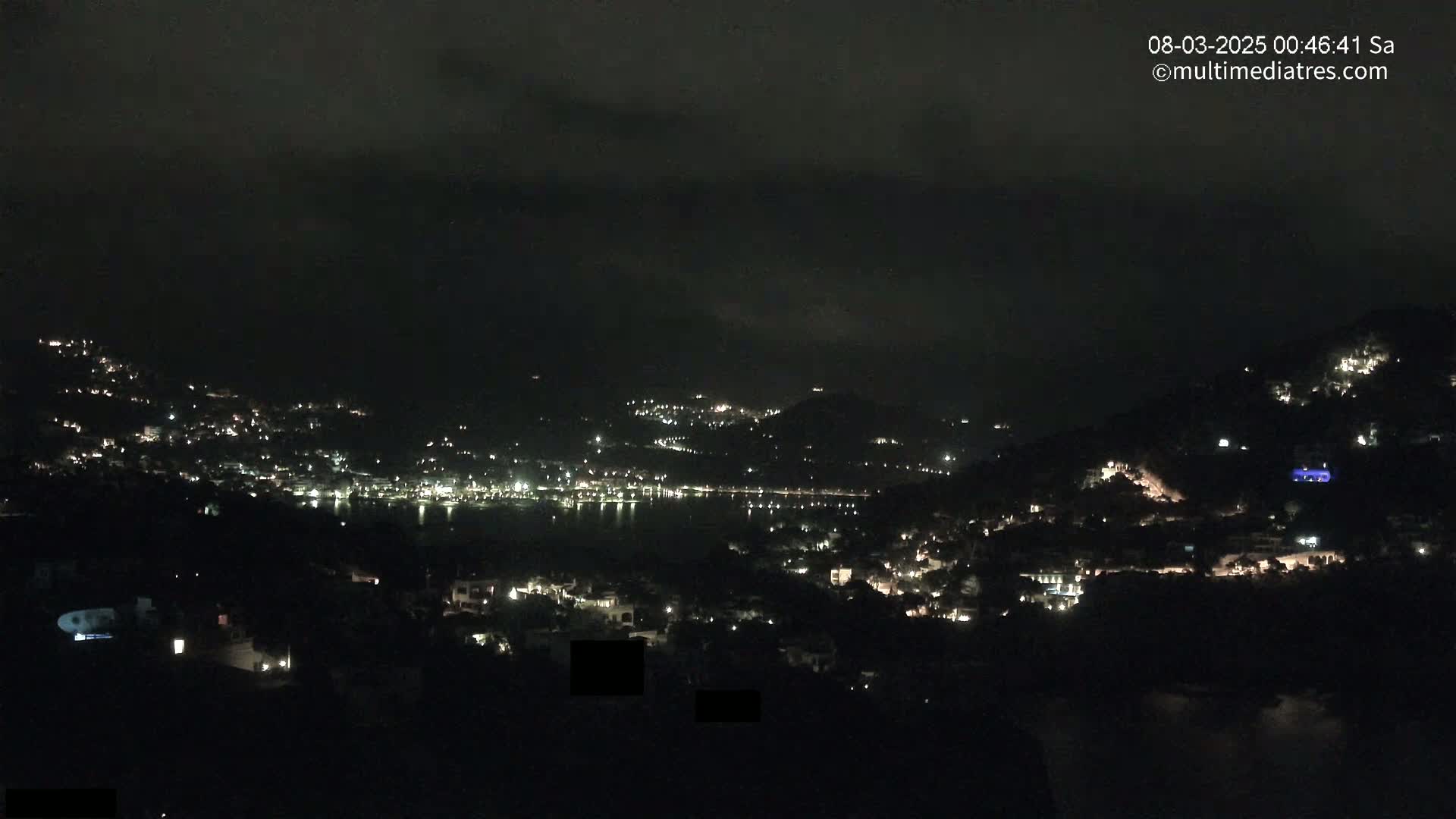 A nighttime view of a coastal town with scattered lights on hillsides surrounding a dark body of water under a cloudy sky.