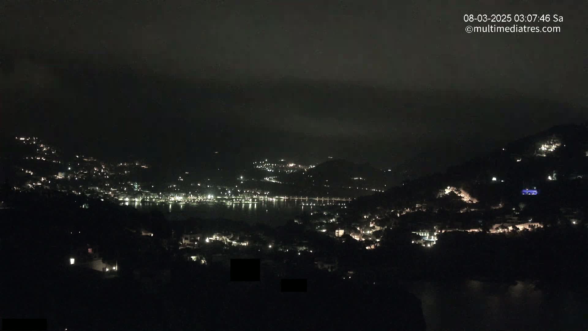 A nighttime view of a coastal town nestled in hills, with scattered lights visible and a body of water in the distance under a dark, cloudy sky.