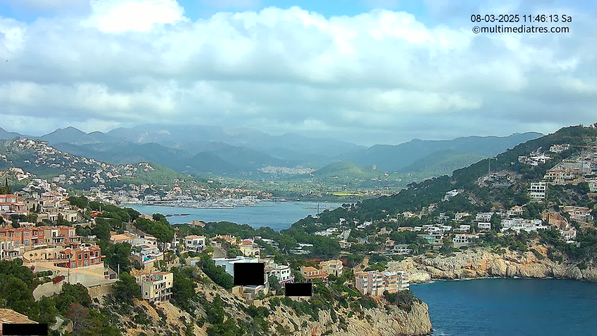 A coastal town nestled on a hillside overlooking a calm bay filled with boats, backed by mountains under a partly cloudy sky.