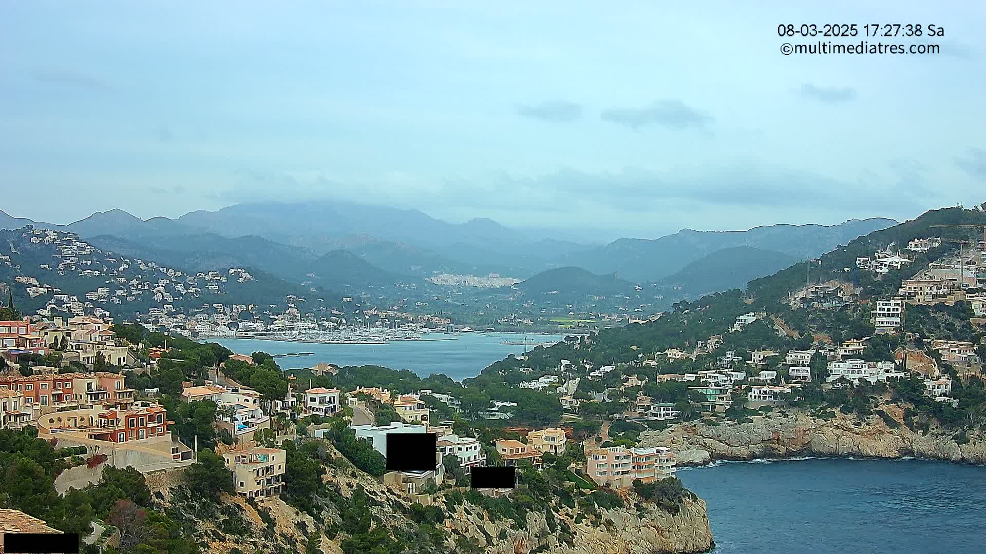 A coastal town nestled on a hillside overlooking a calm bay, with mountains in the background under a mostly cloudy sky.