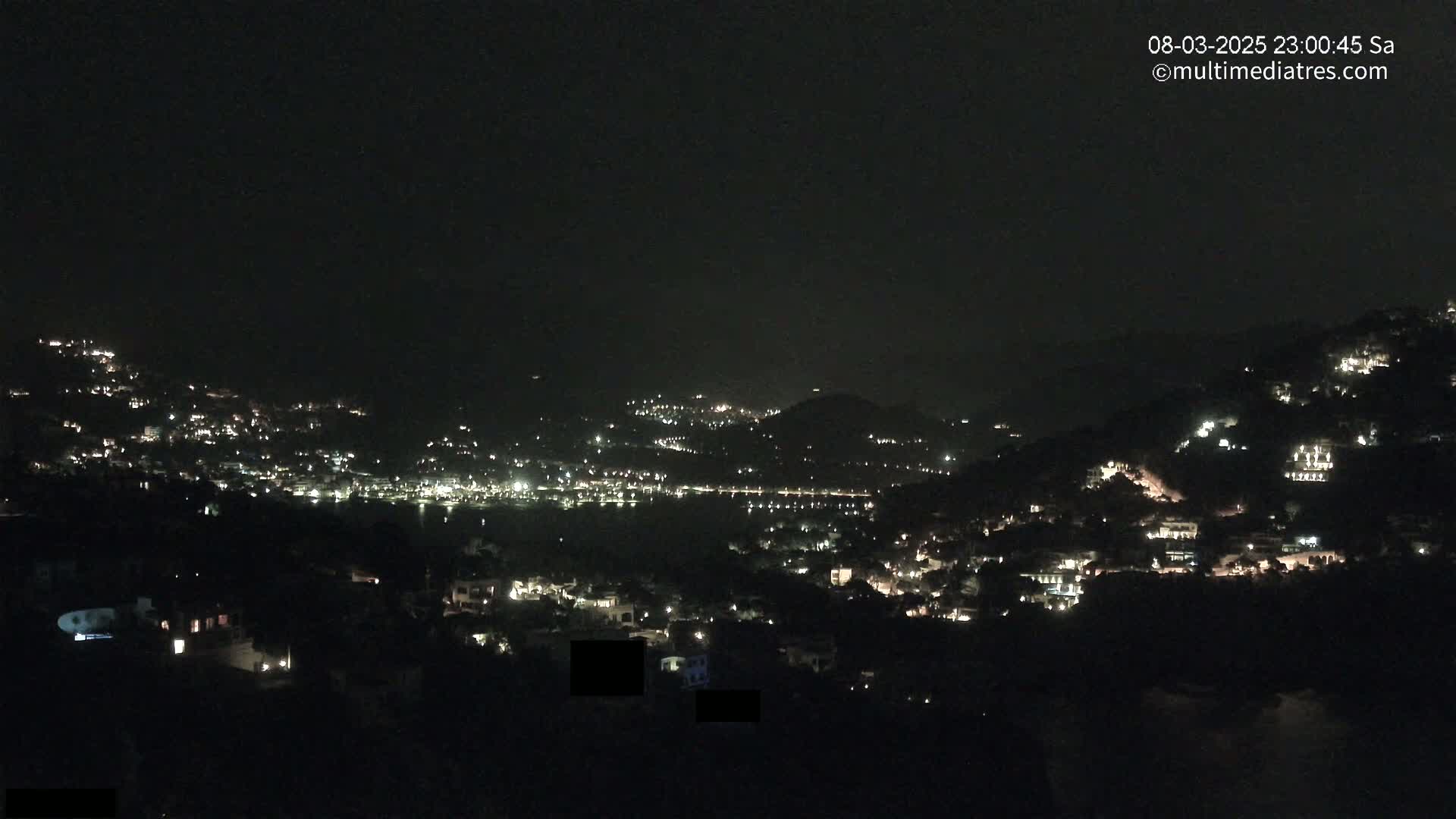 A nighttime view of a town nestled on hillsides surrounding a body of water, illuminated by scattered lights under a clear, dark sky.