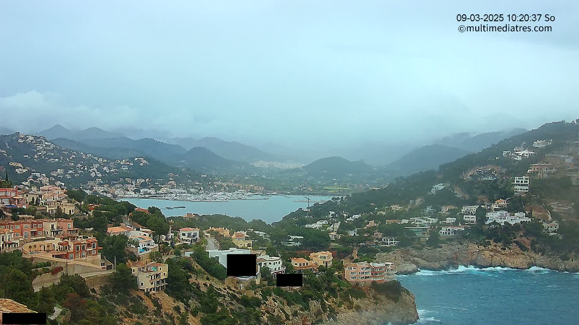 A coastal town nestled into hills overlooking a calm bay with numerous boats, under an overcast sky.