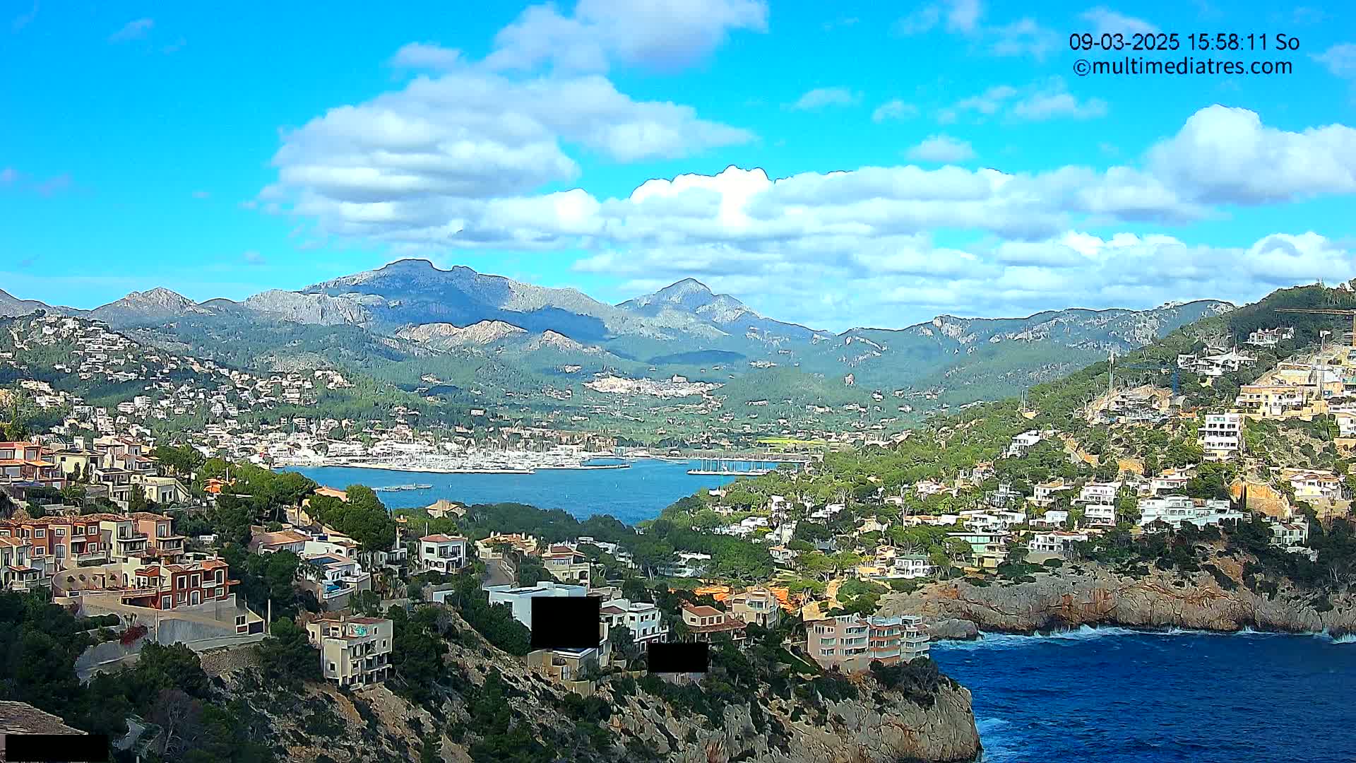A coastal town nestled between a mountain range and a blue bay under a partly cloudy sky.