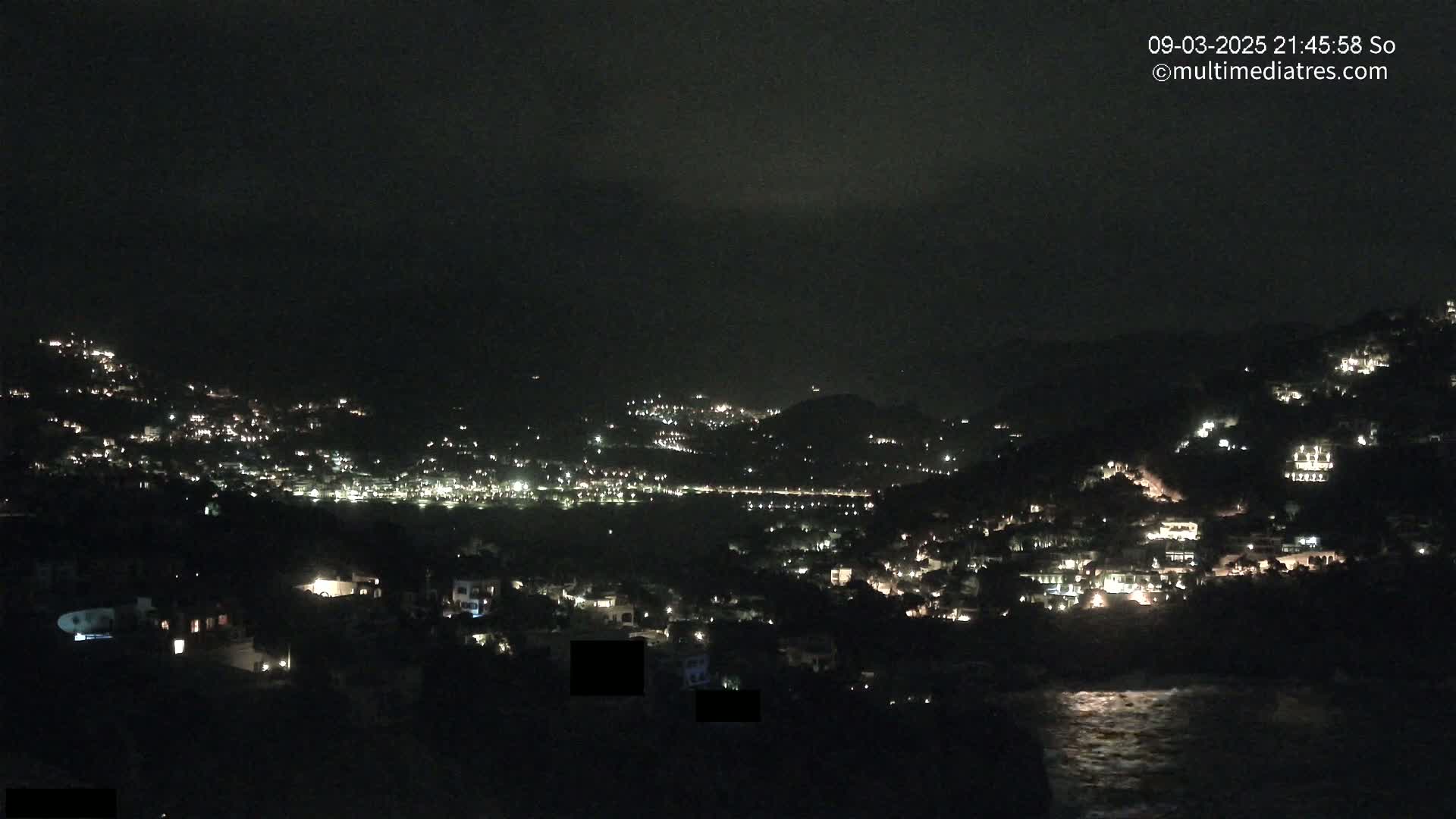 A nighttime view of a coastal town nestled on hillsides, illuminated by scattered lights, under a dark, cloudy sky.