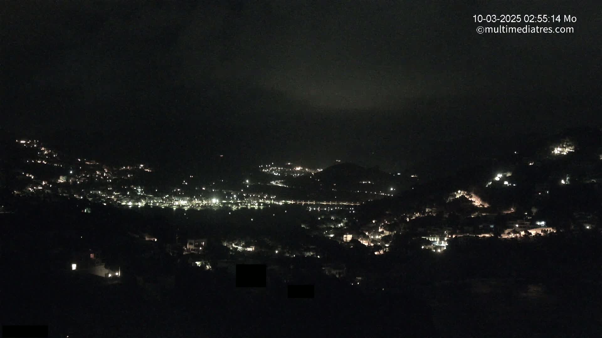 A nighttime view shows a hillside town illuminated by scattered lights under a dark, possibly cloudy sky.