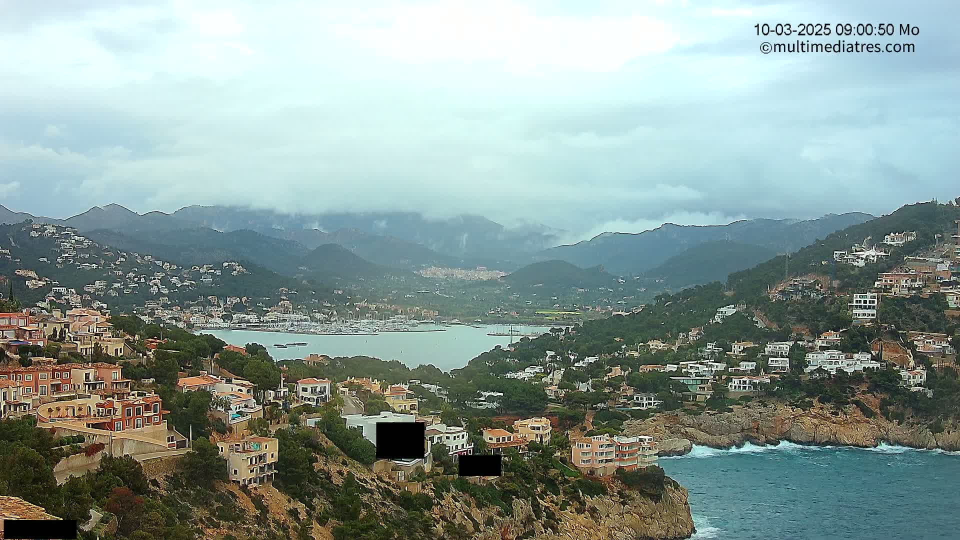 A coastal town nestled in a mountainous region with an overcast sky, overlooking a calm bay filled with boats.