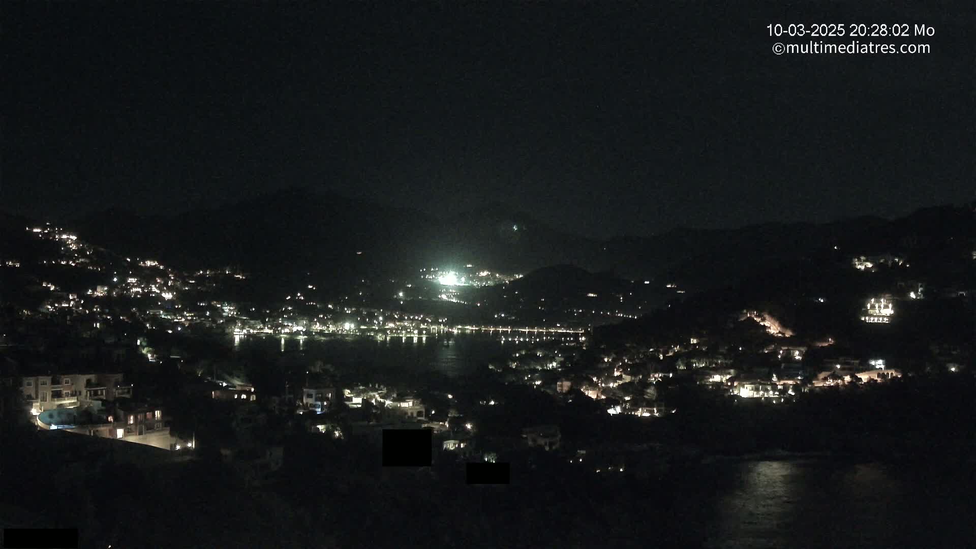 A nighttime view of a coastal town illuminated by lights, nestled between dark hills and a calm body of water under a clear, dark sky.