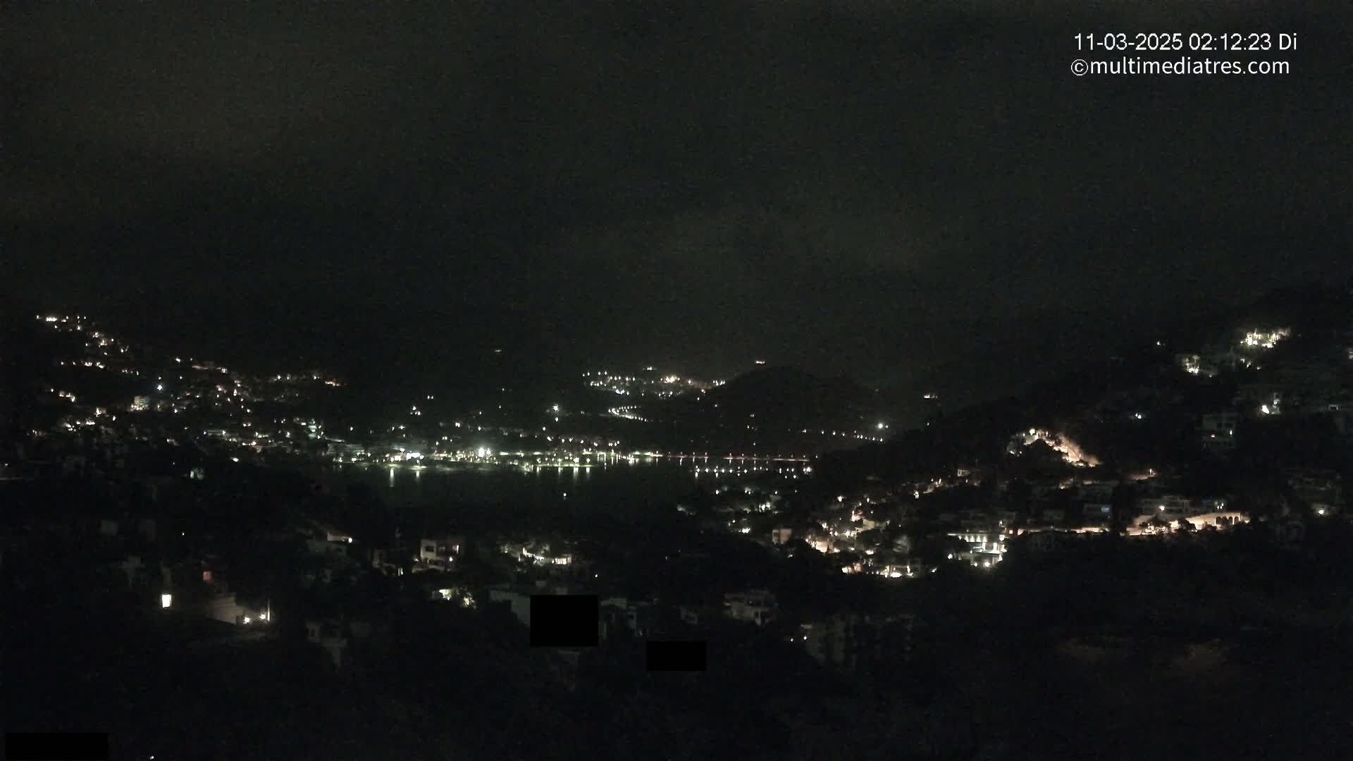 A nighttime view of a coastal town, with hills dotted with lights, overlooking a dark body of water under a mostly clear sky.