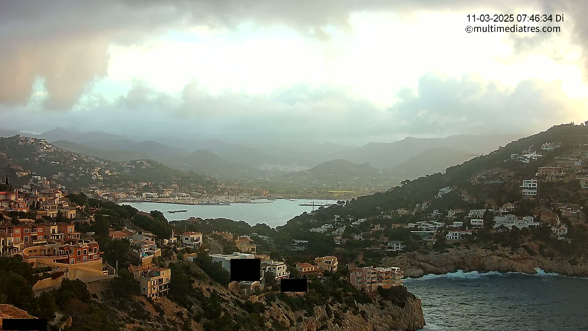 A coastal town nestled on hillsides overlooking a calm bay with many boats, under a mostly cloudy sky.