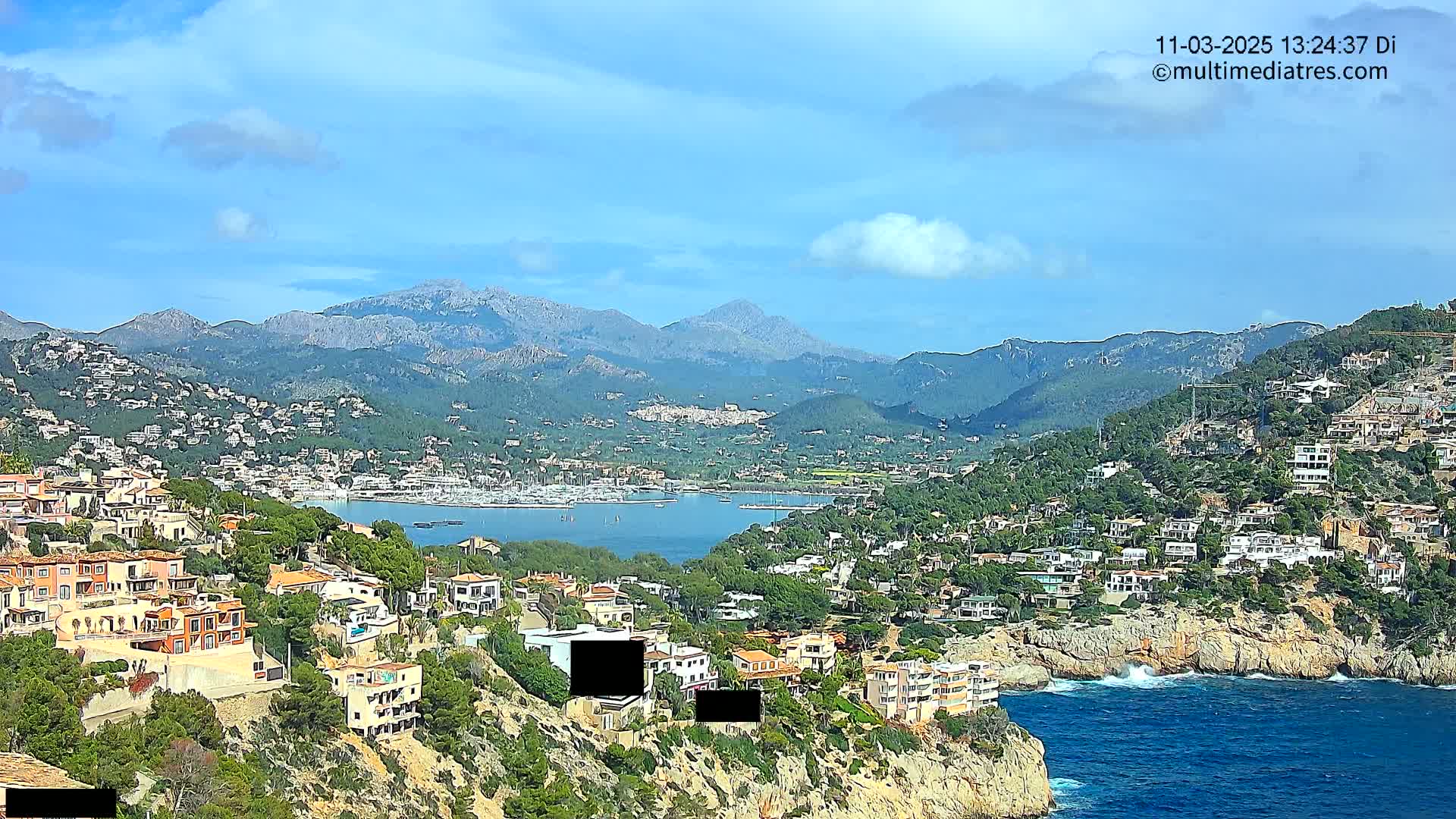 A coastal town nestled on a hillside overlooking a calm bay with numerous boats, backed by mountains under a mostly clear, bright sky.