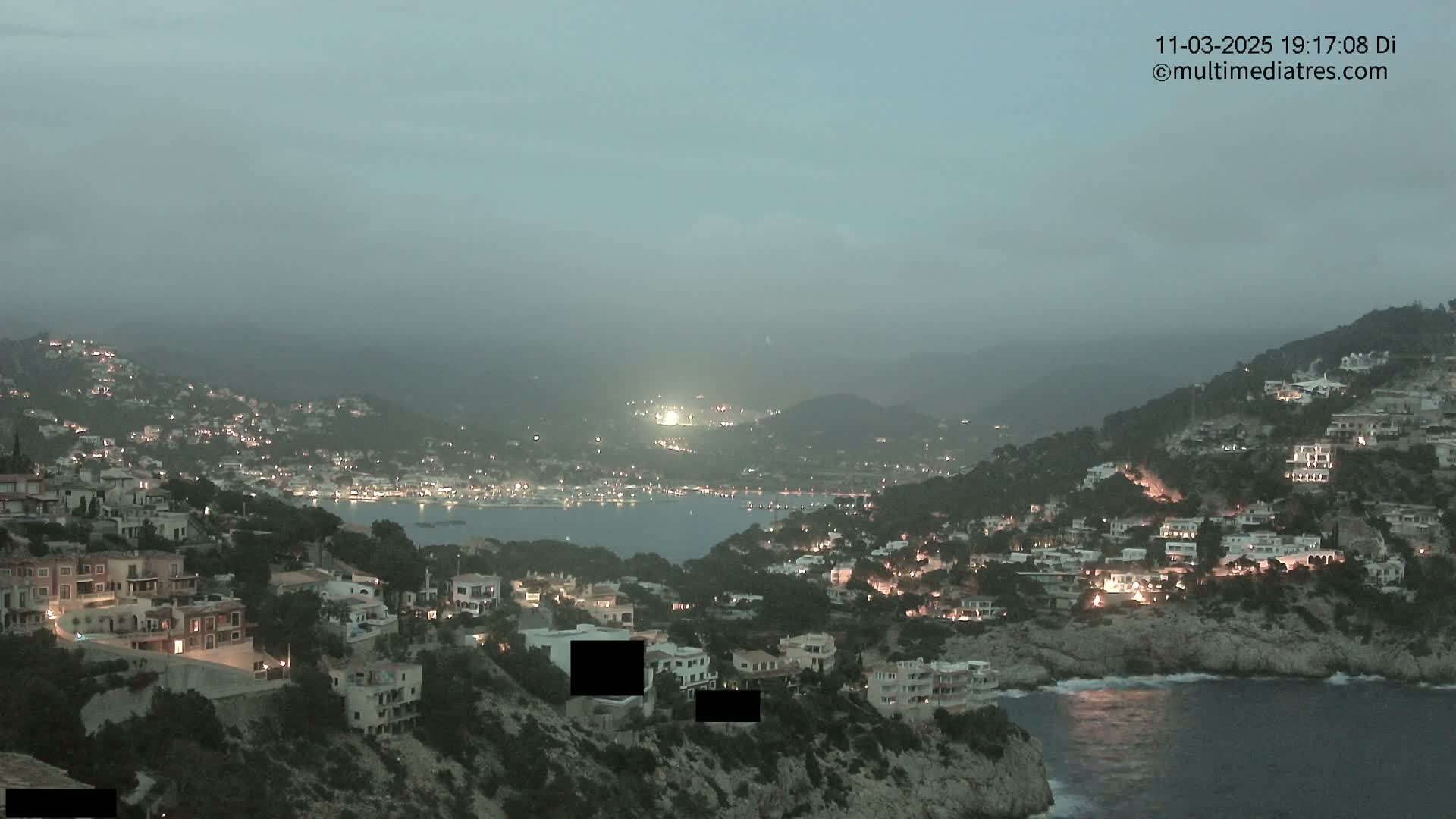 A coastal town at twilight, under a hazy sky, is illuminated by lights, nestled between hillsides and a dark body of water.
