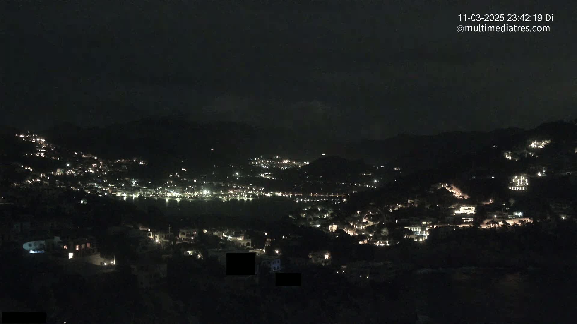 A nighttime view of a coastal town illuminated by scattered lights, nestled between dark hills under a clear, dark sky.