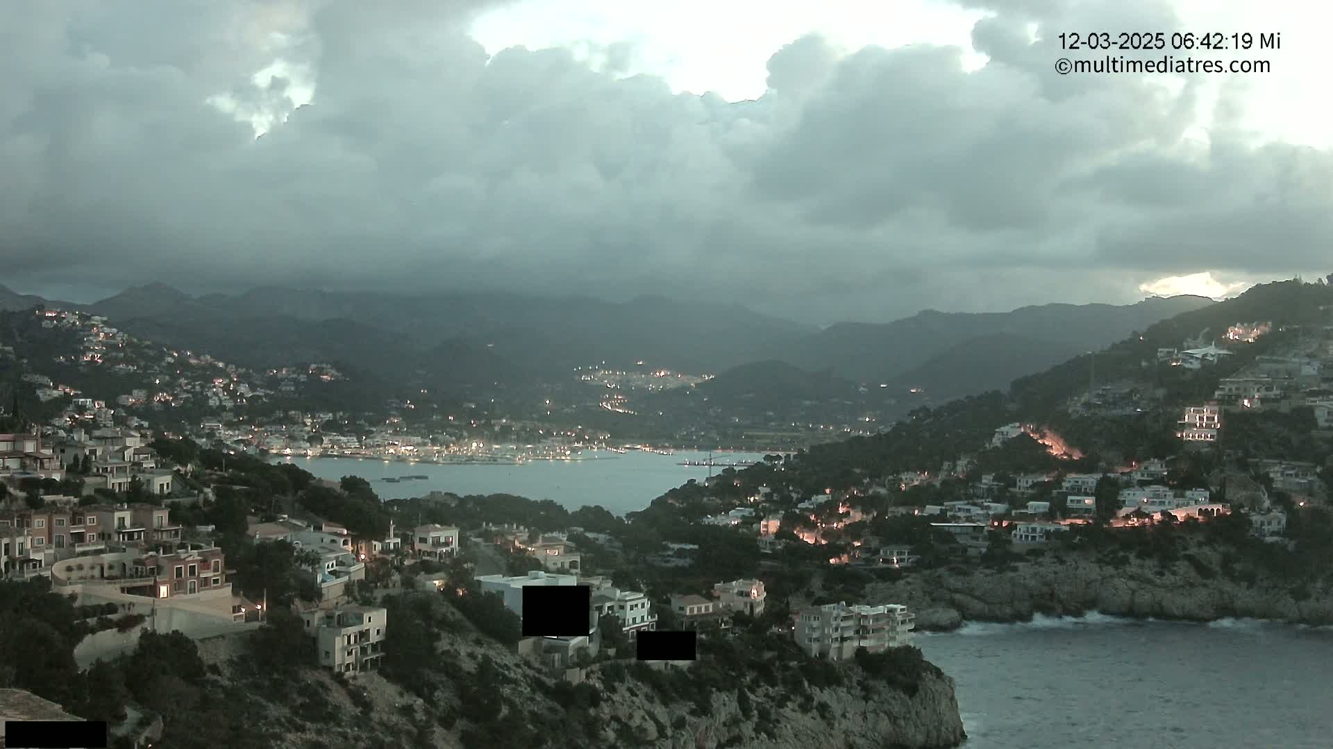 A coastal town at dusk, nestled between hills under a cloudy sky, with lights illuminating buildings and a calm body of water below.