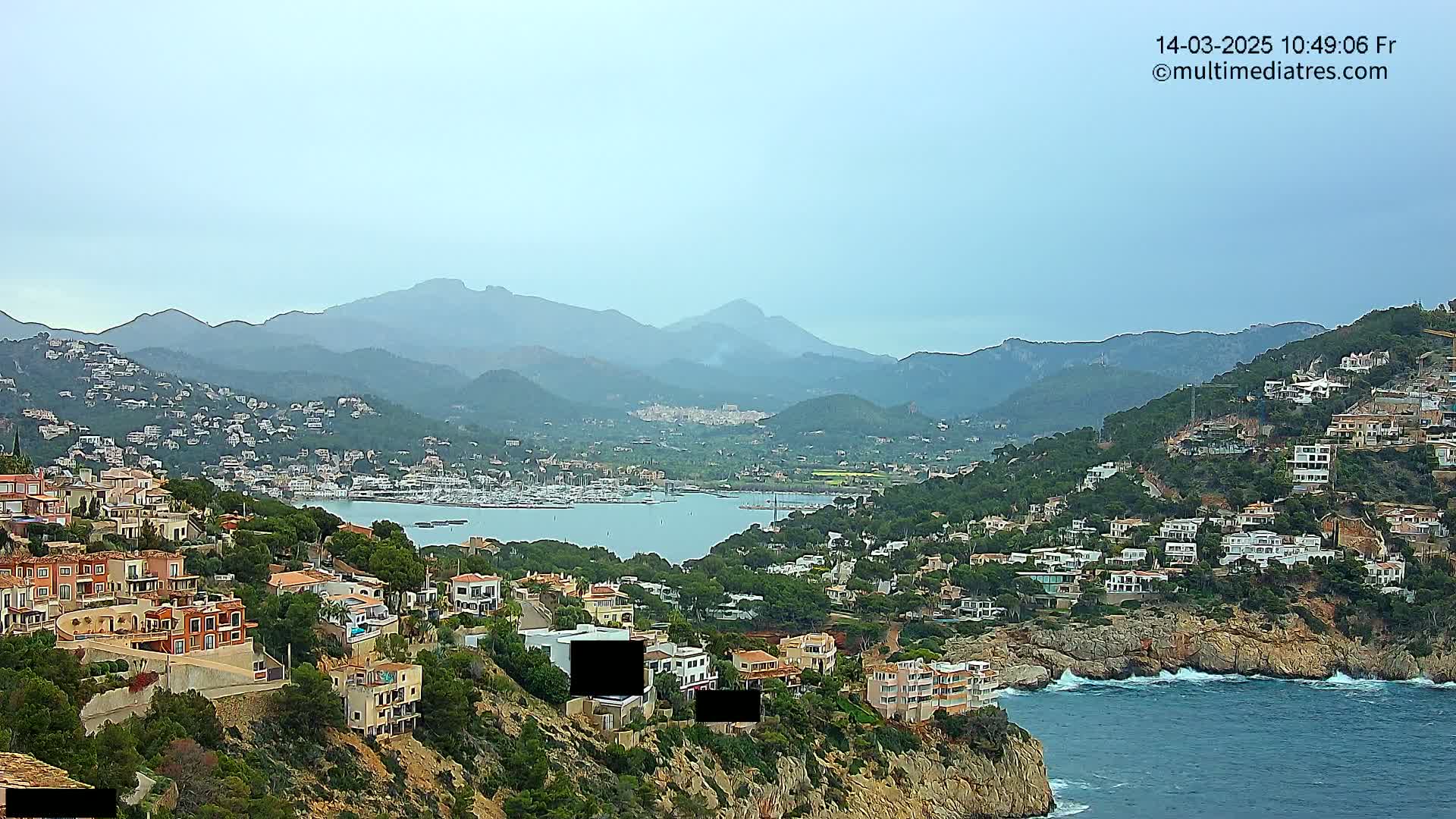 A coastal town nestled on a hillside overlooking a calm bay filled with boats, with hazy mountains in the background under an overcast sky.