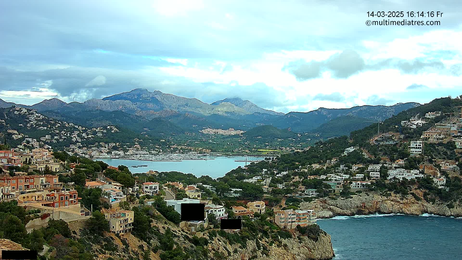 A coastal town nestled into a hillside overlooking a calm bay, with mountains in the background under a partly cloudy sky.