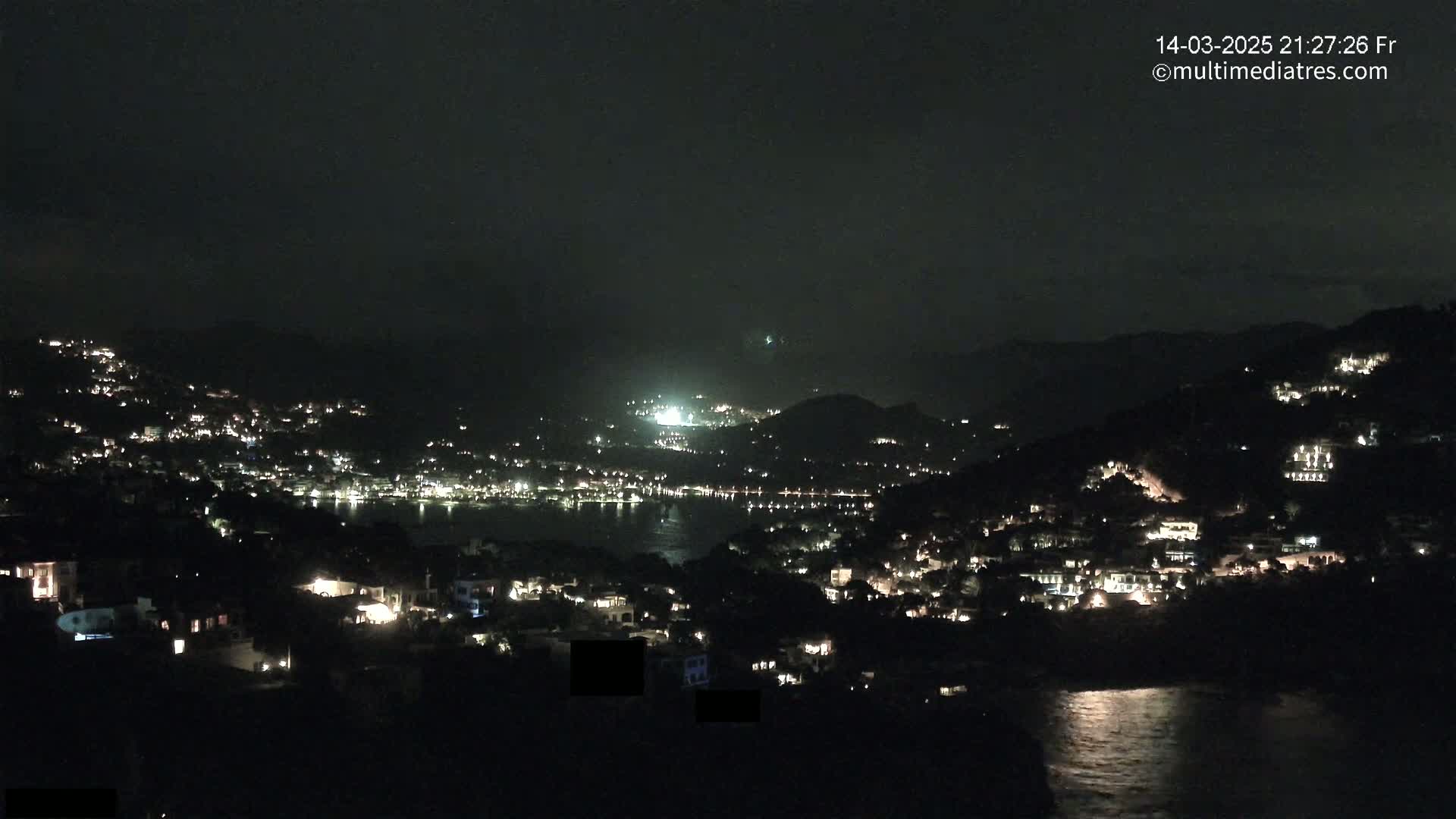 A nighttime view of a coastal town nestled on hillsides, with scattered lights illuminating buildings and a dark body of water reflecting some light.