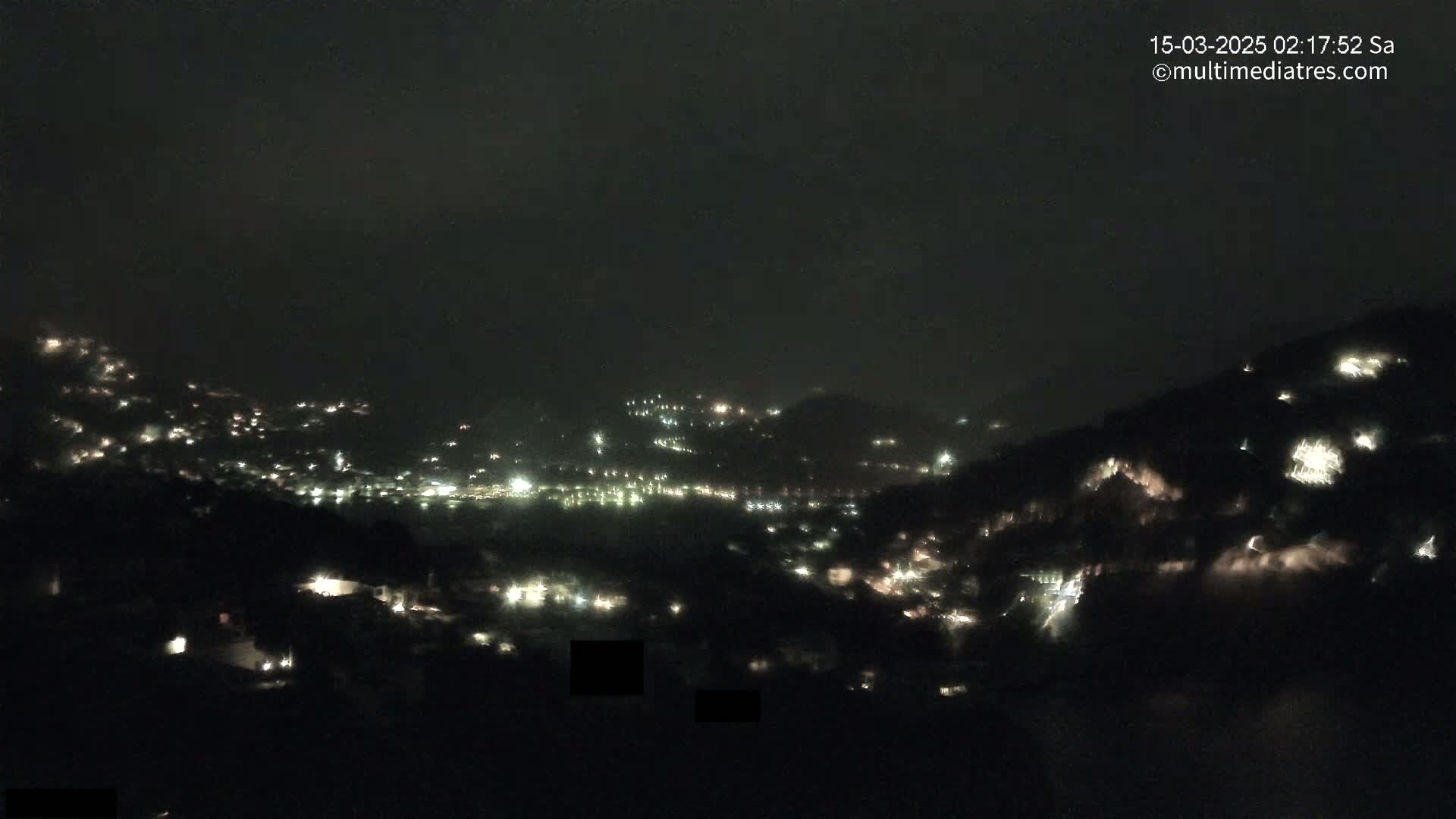 A nighttime view of a hilly town speckled with scattered lights under a dark, cloudy sky.