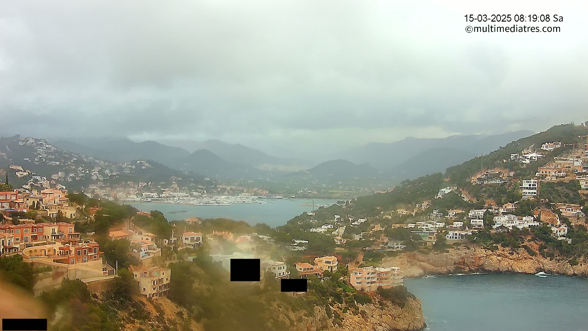 A coastal town nestled on a hillside overlooks a calm bay filled with boats under an overcast sky.