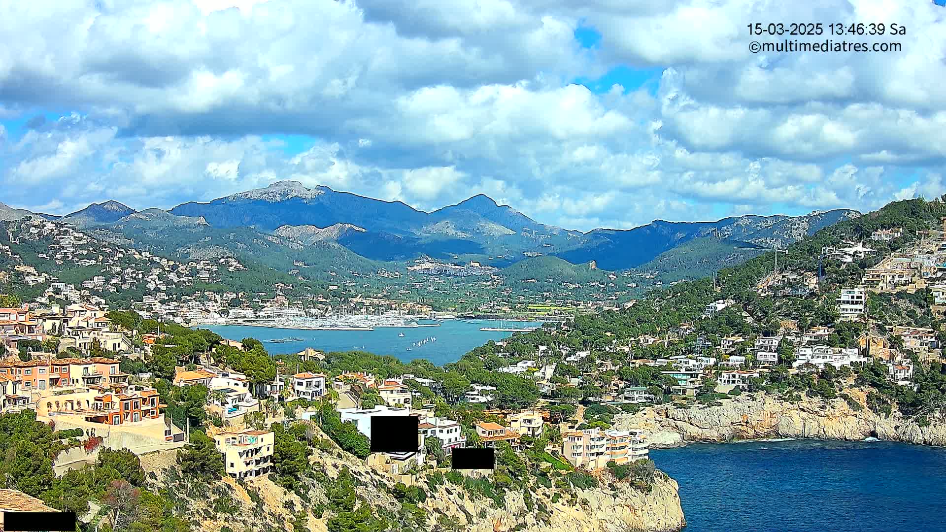 A coastal town nestled among hills, with a harbor filled with boats and a partly cloudy sky.